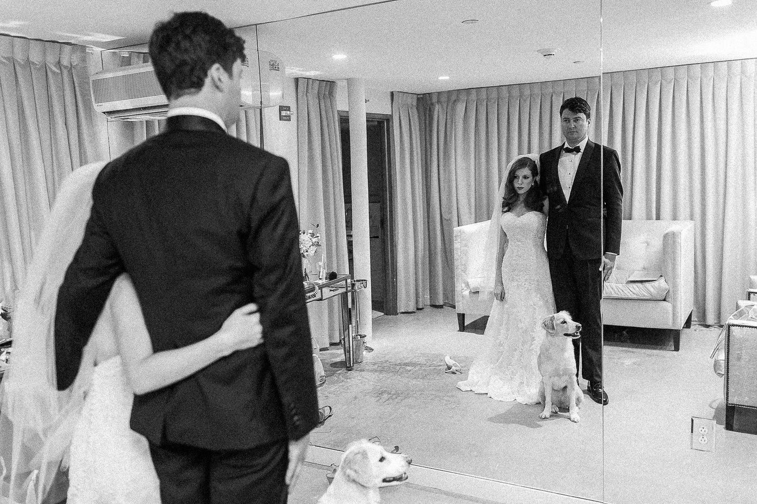 Black and white wedding photo of couple viewing their reflection in a large mirror with a white dog nearby.