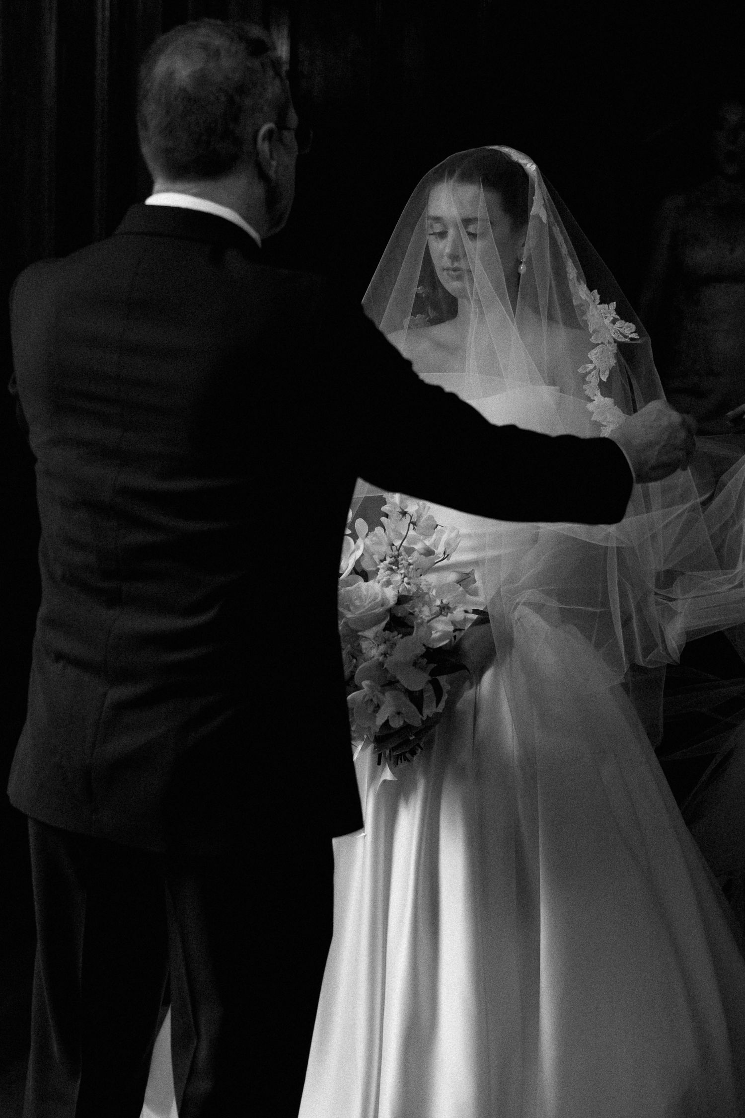 A black and white wedding photograph showing a bride in a traditional white gown and veil holding a floral bouquet.