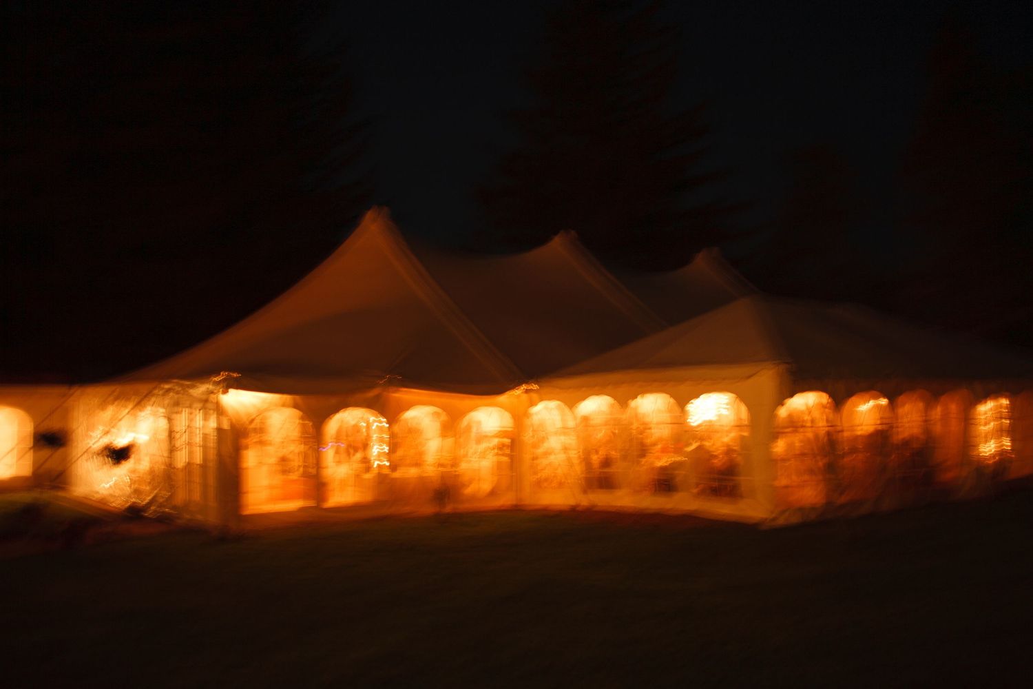 Illuminated white event tent glowing warmly against a dark night sky at an outdoor wedding reception.