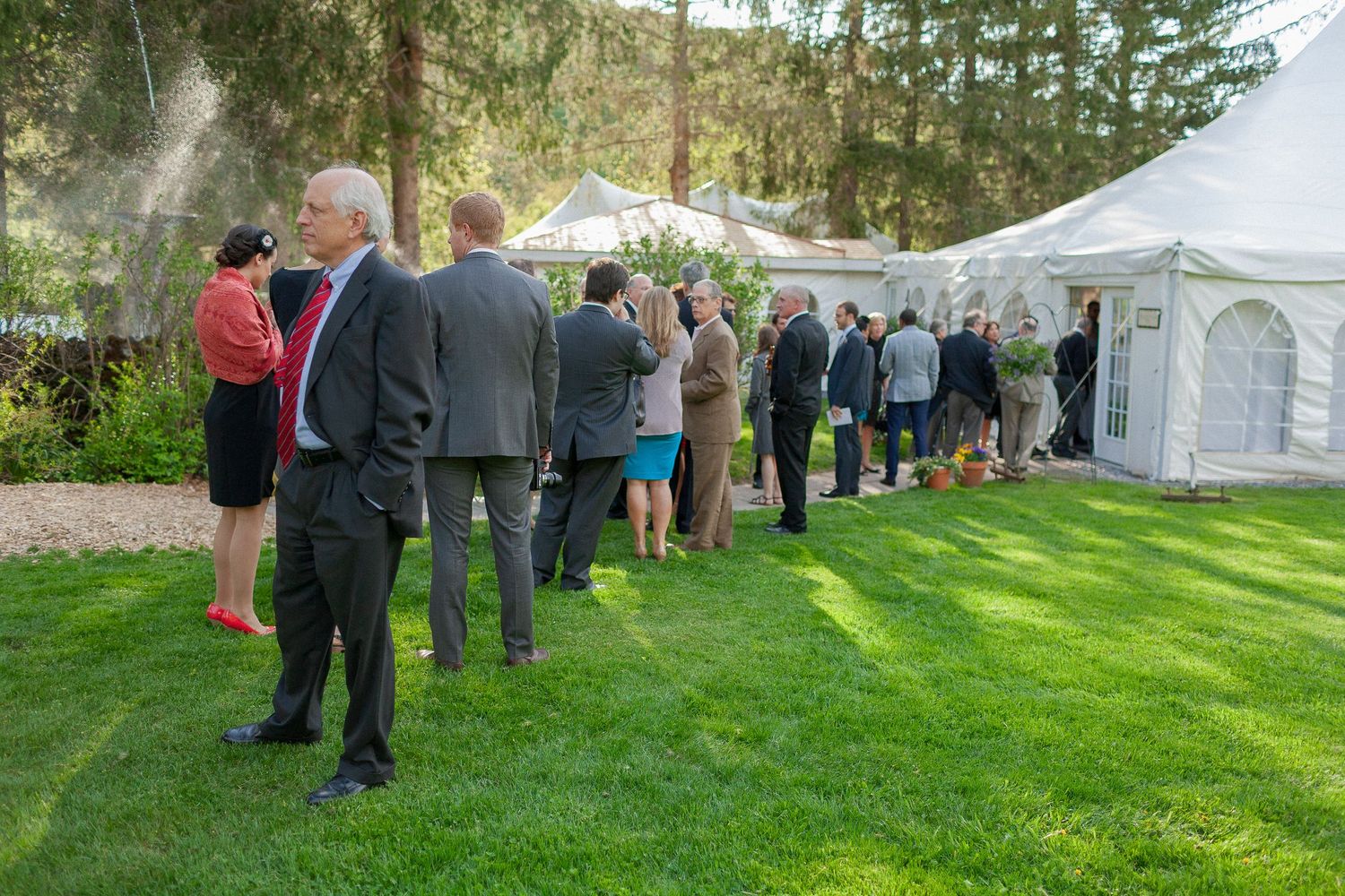 Guests in formal attire line up outside a white event tent on a lush green lawn at an outdoor garden reception.