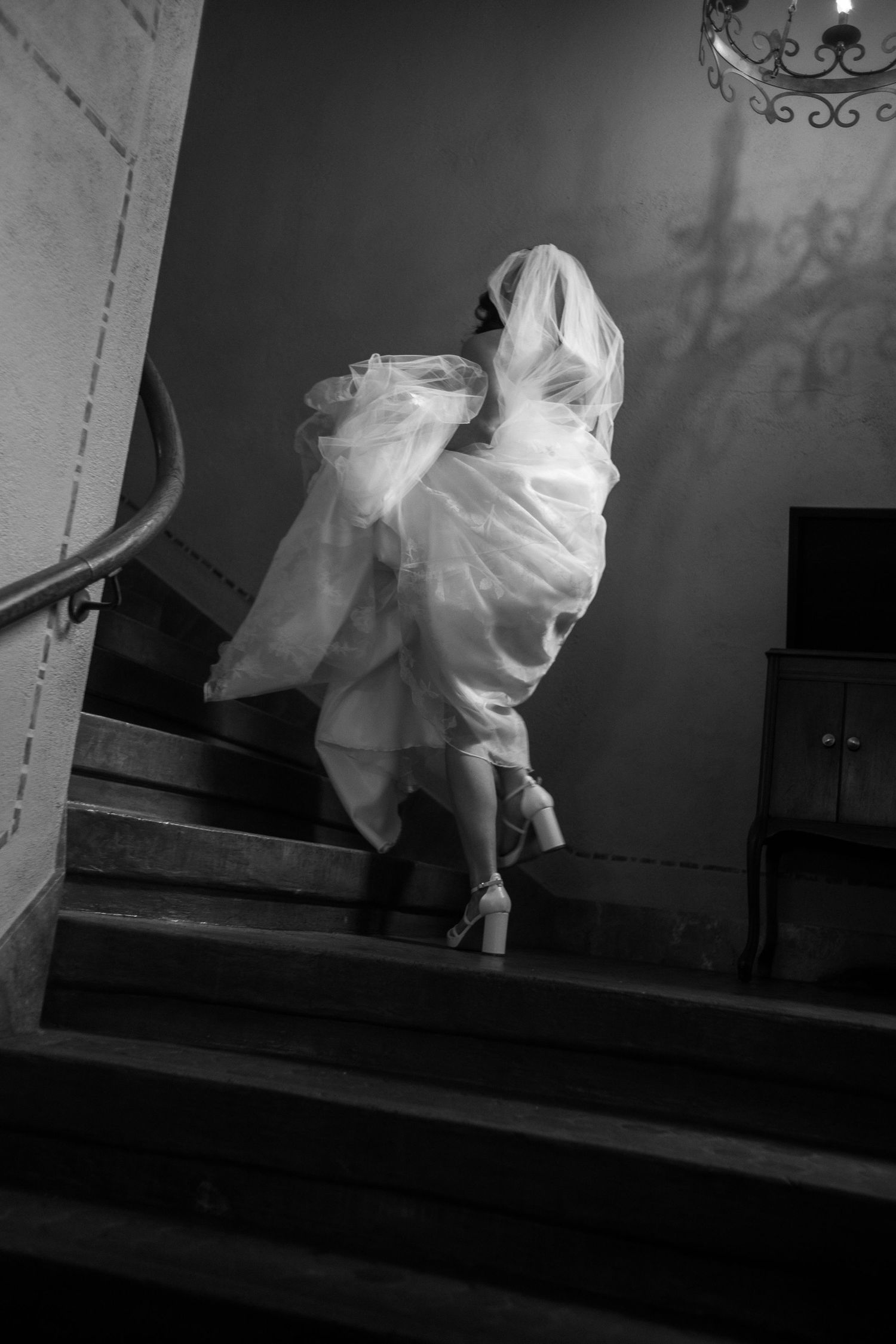 Black and white photo of a bride in a flowing gown and veil ascending a grand staircase in heels.