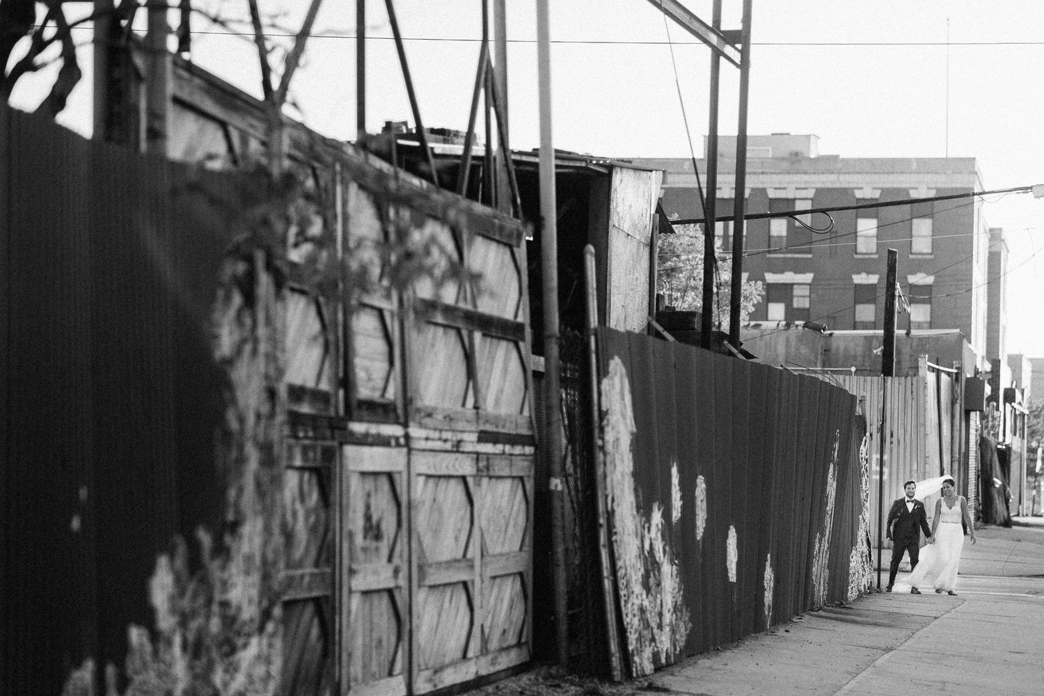 Black and white photo of a weathered urban alleyway with rusty metal fences and a brick building in the background.
