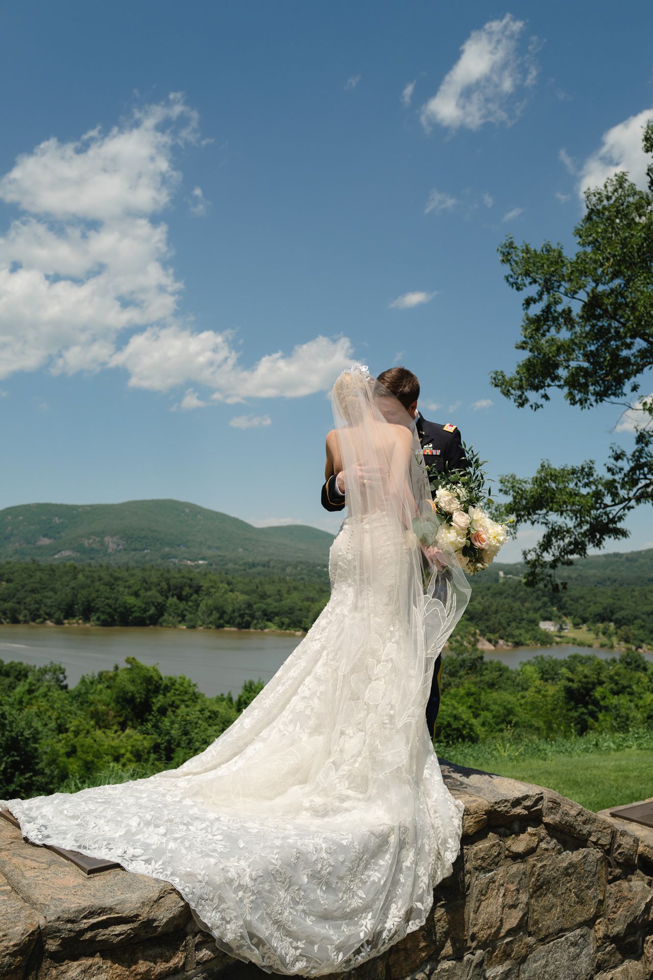 A wedding dress with lace train flows dramatically against a mountain landscape backdrop.