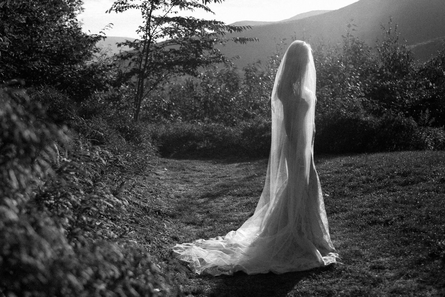 Black and white photo of a bride in a flowing white wedding dress and veil standing on a rural path.