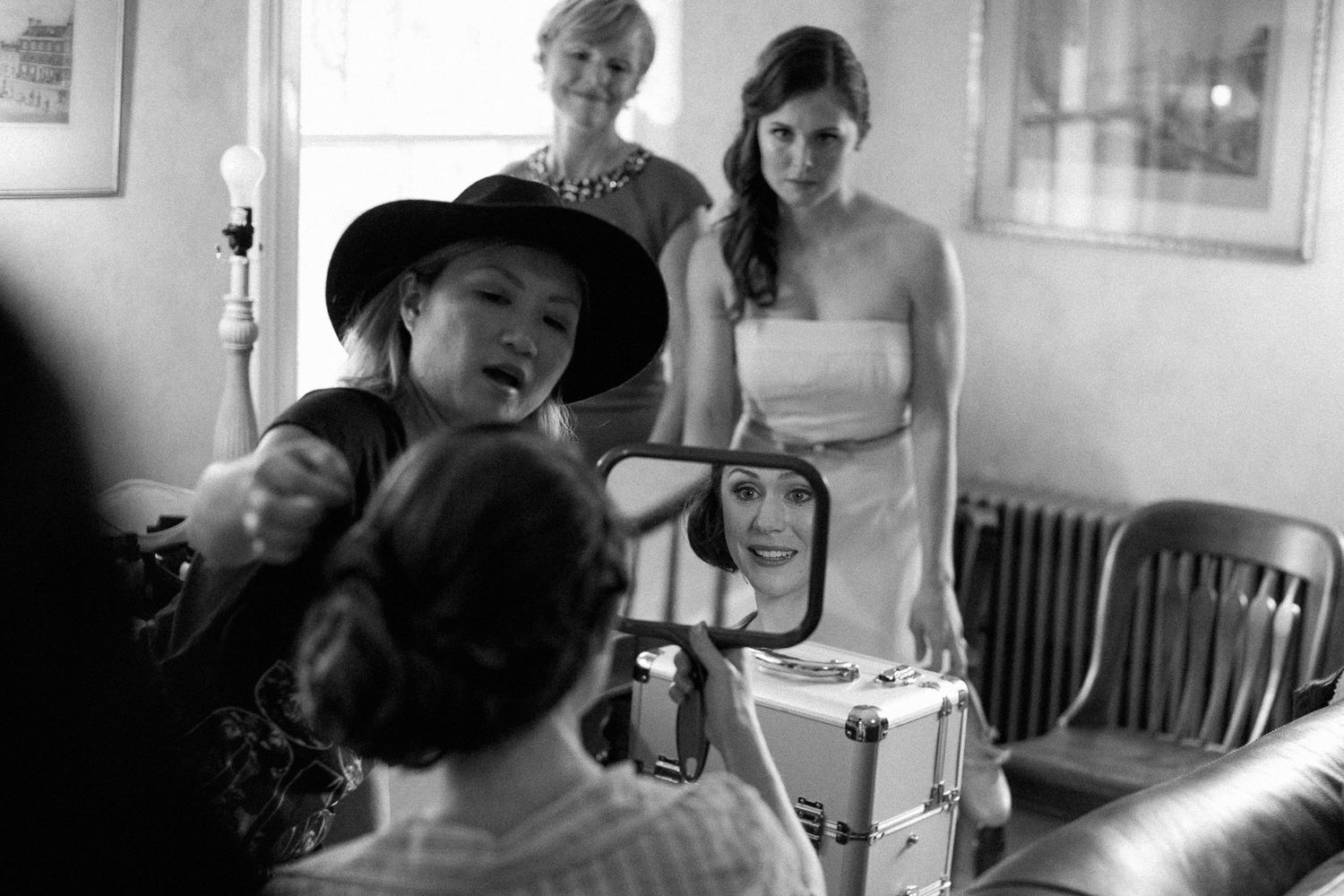 Black and white photo of a bride getting ready, with a makeup artist holding a mirror and guests watching.