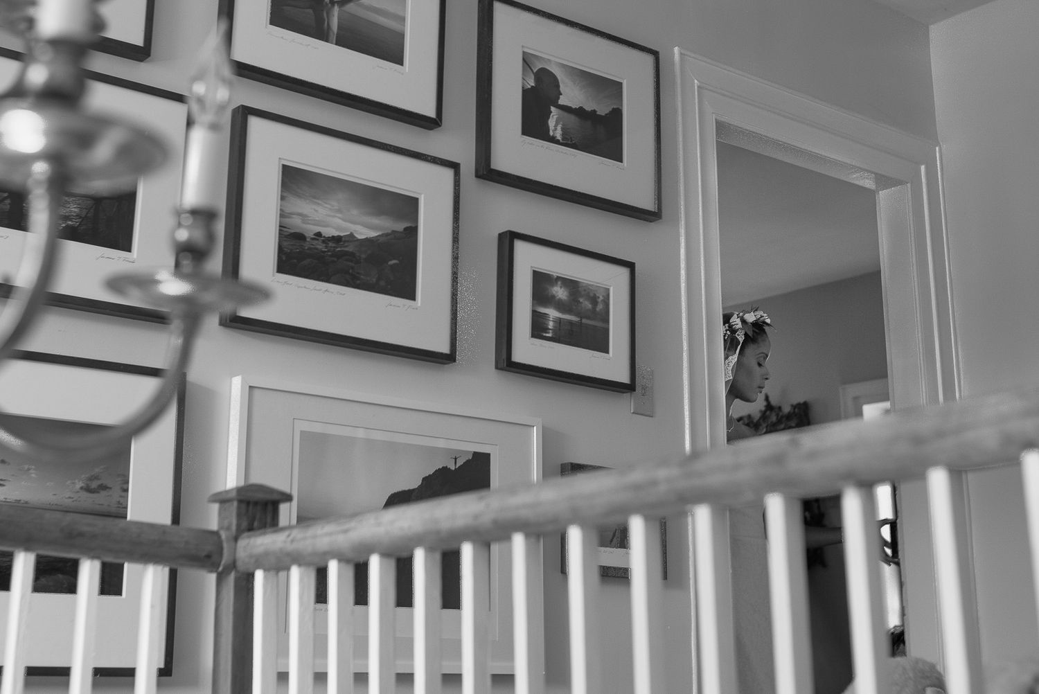 Black and white photo of a staircase with white railings and a gallery wall of framed pictures in a home interior.