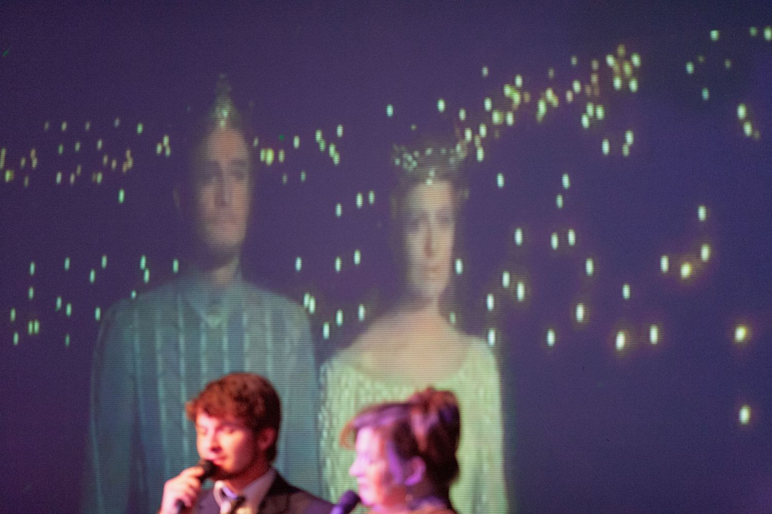 Two performers on stage with microphones, backed by a projected image of a couple in formal attire on a purple backdrop.