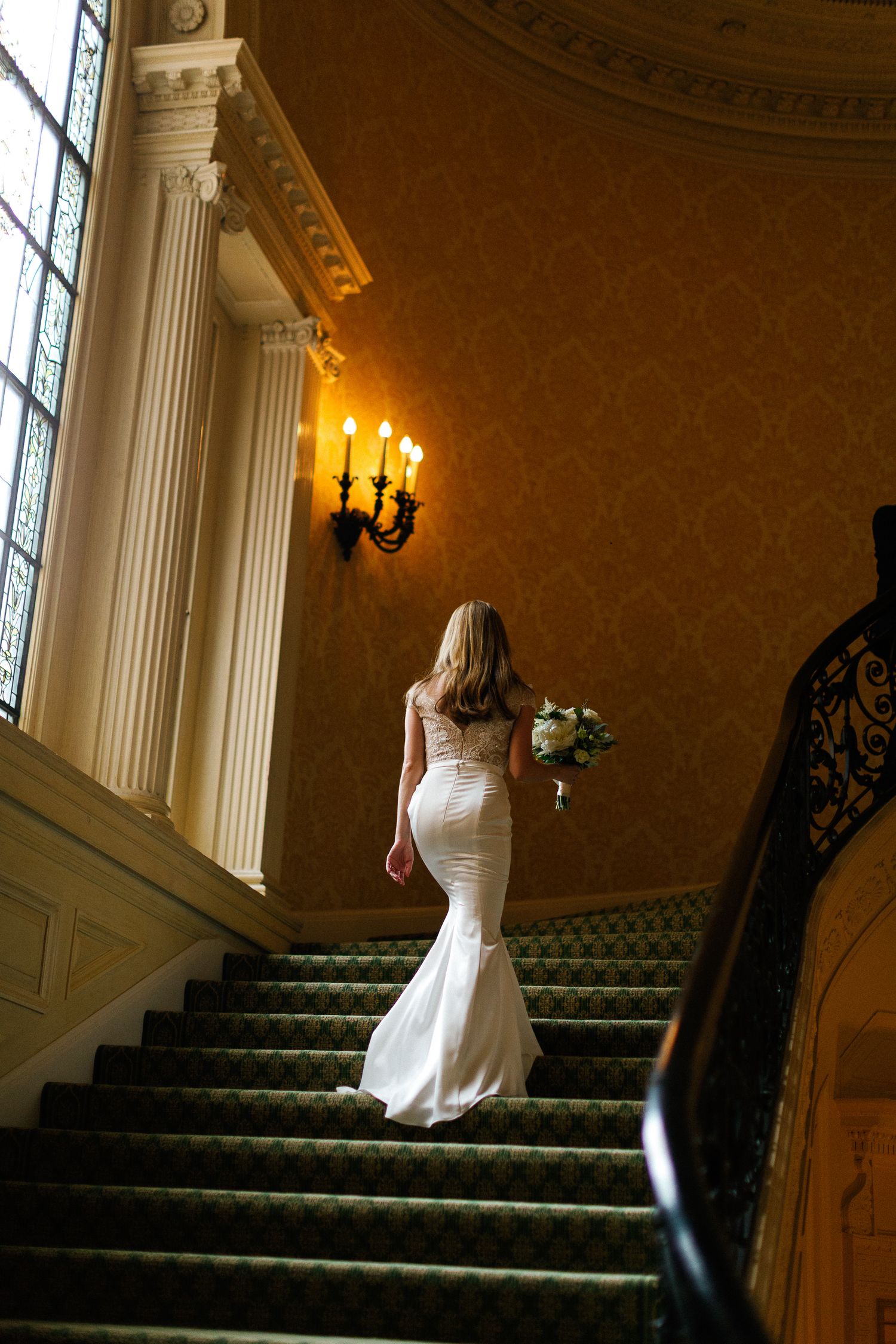 Bride in white mermaid gown ascending grand staircase holding bouquet in elegant venue with candle wall sconces.