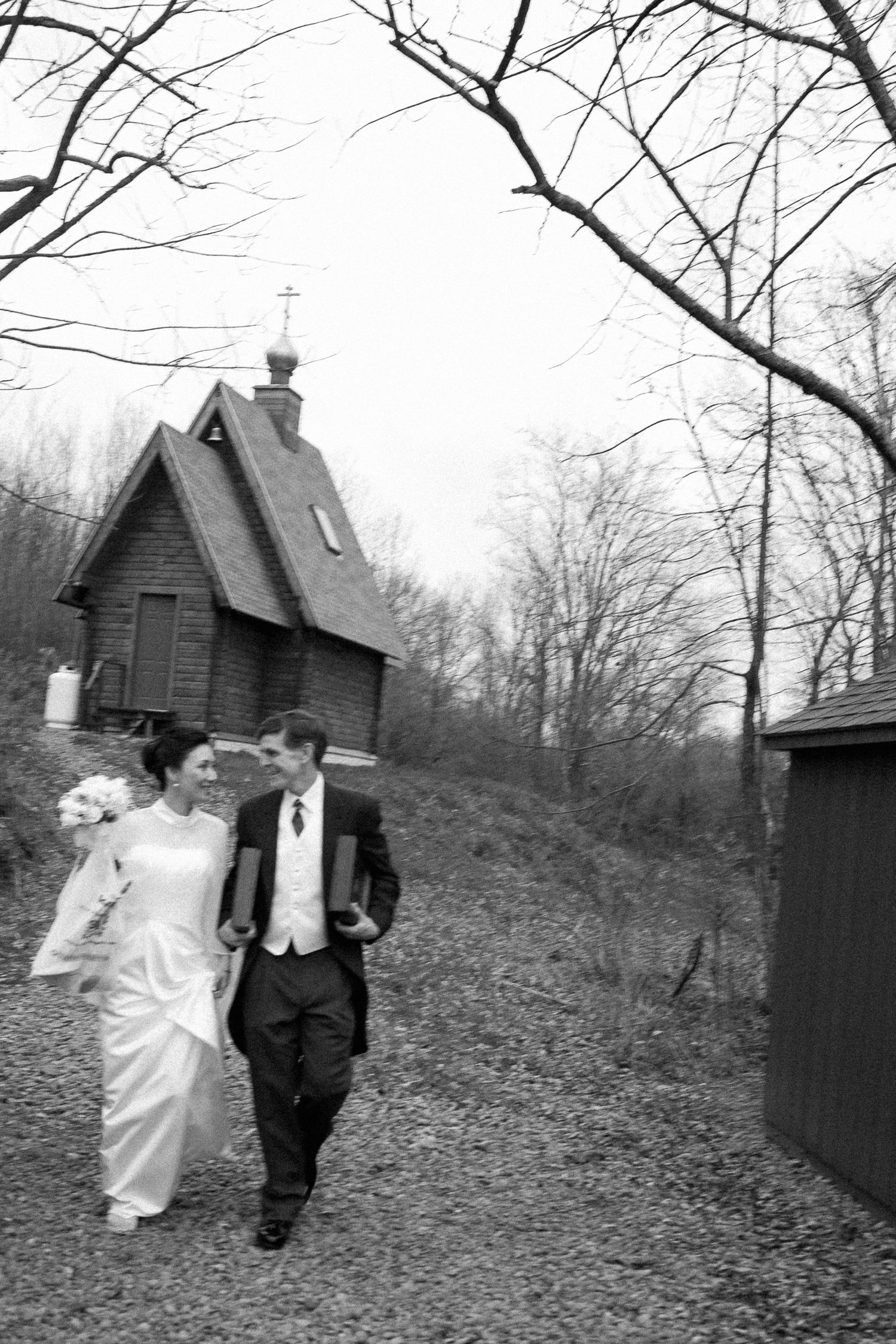 Black and white photo of a bride and groom walking hand in hand near a small rustic chapel surrounded by bare trees.