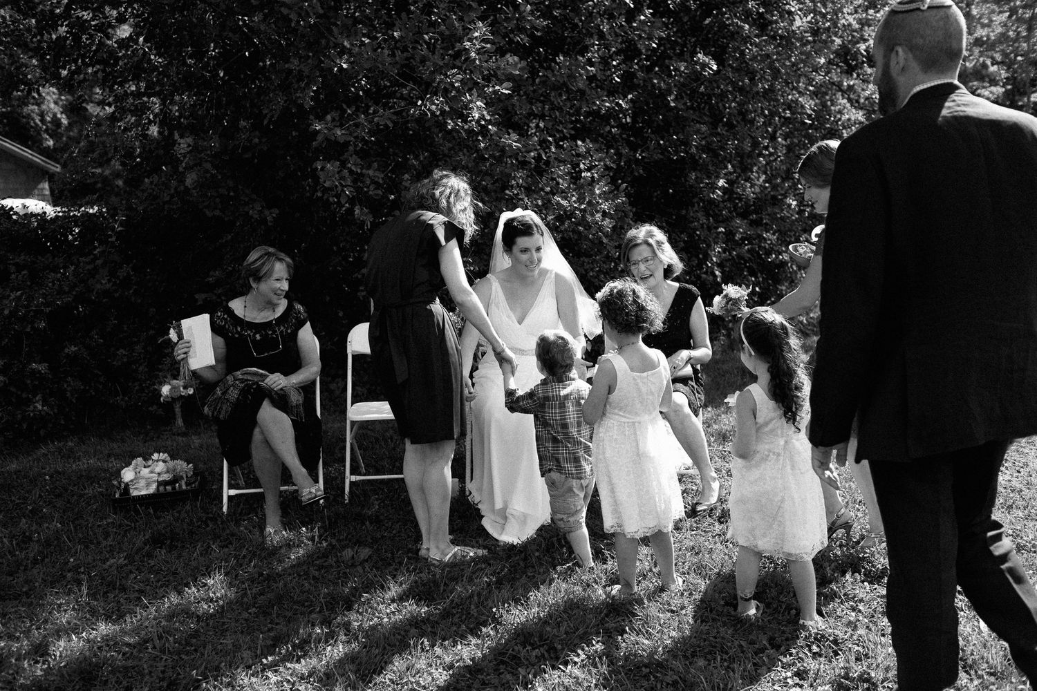 Black and white photo of a bride in a wedding gown greeting children and guests outdoors during a garden ceremony.
