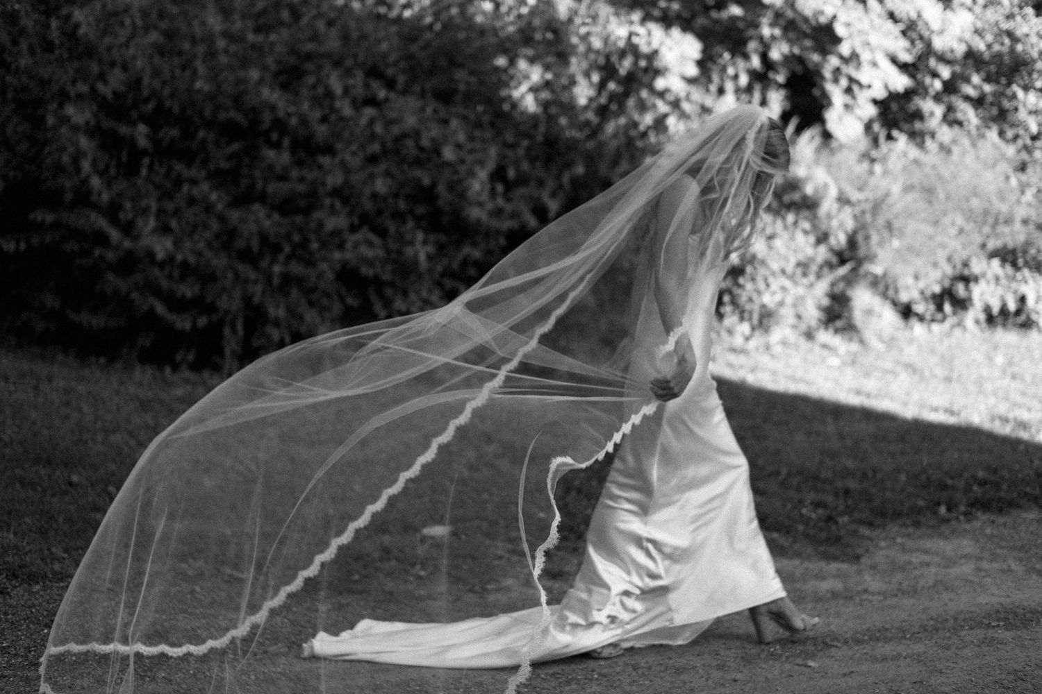 A flowing bridal veil catches the wind as someone in a white dress runs through a garden setting in black and white.