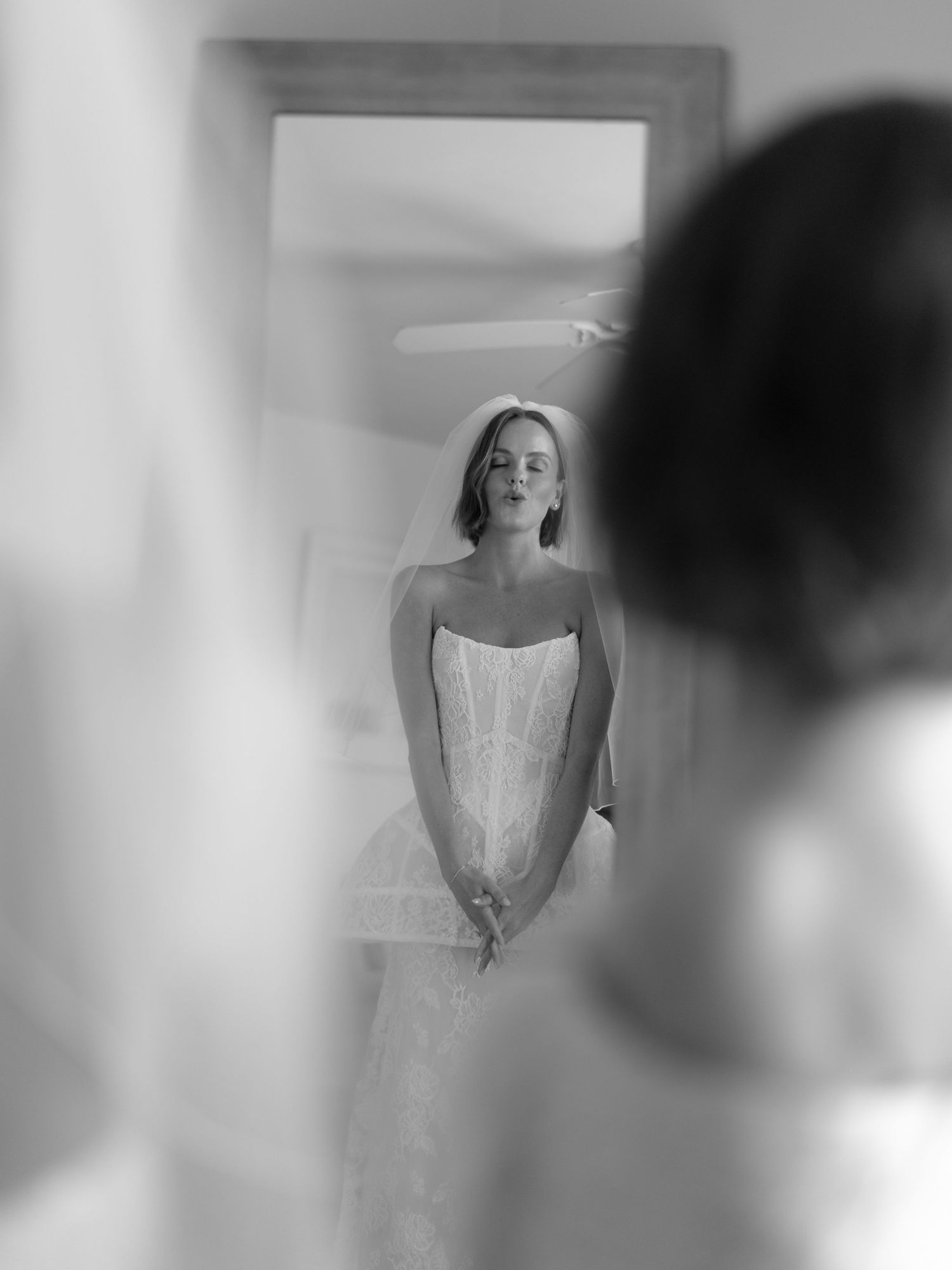 A black and white artistic photograph of a bride in a strapless wedding dress looking at her reflection in a mirror.