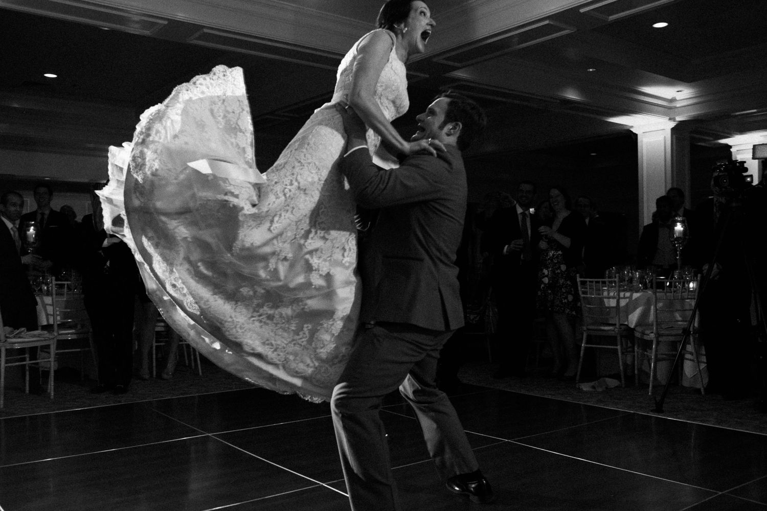 Black and white photo of groom lifting bride in wedding dress during first dance at reception.
