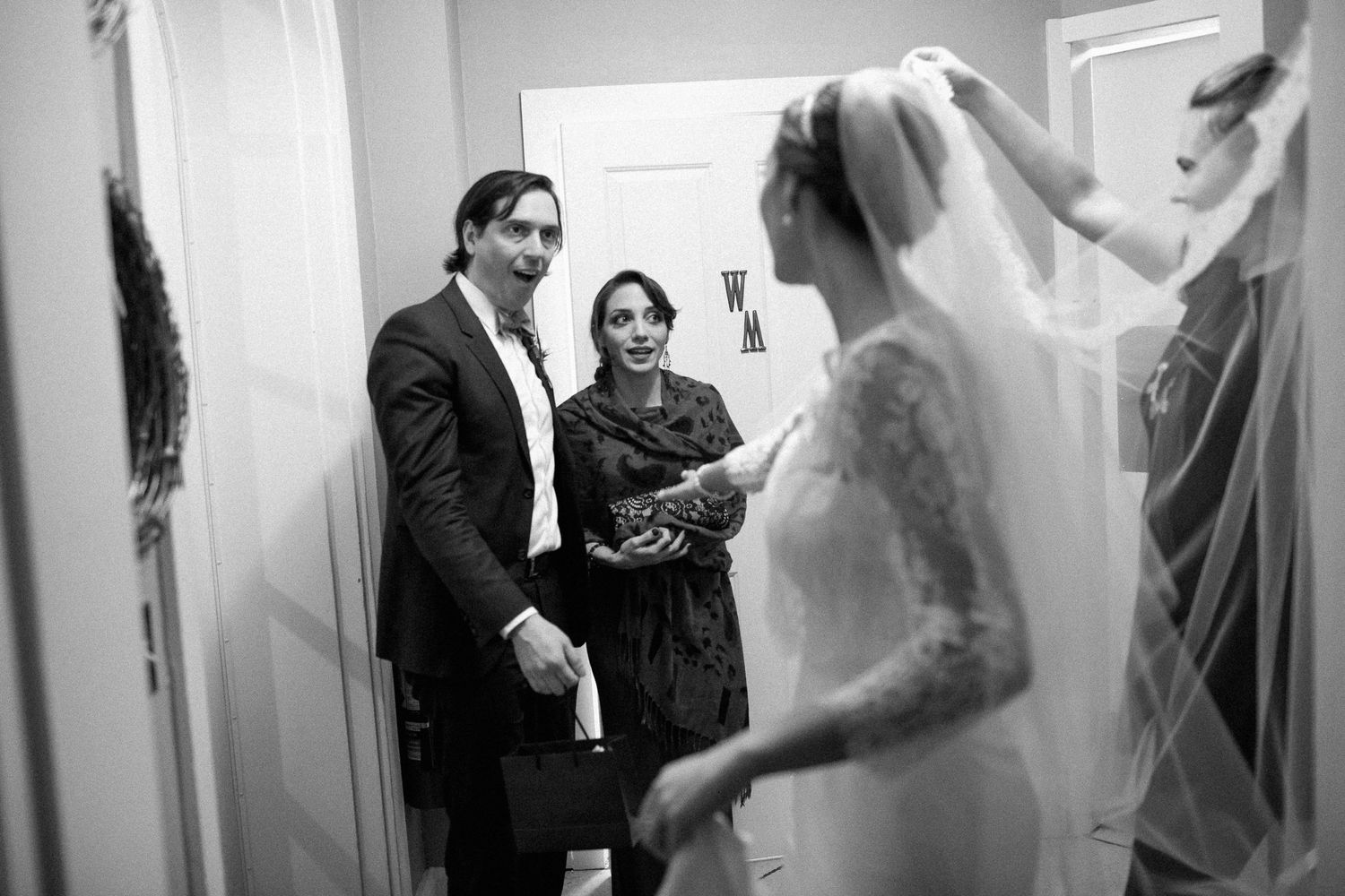 Black and white photo of a bride in lace gown and veil being admired by smiling guests in a hallway.