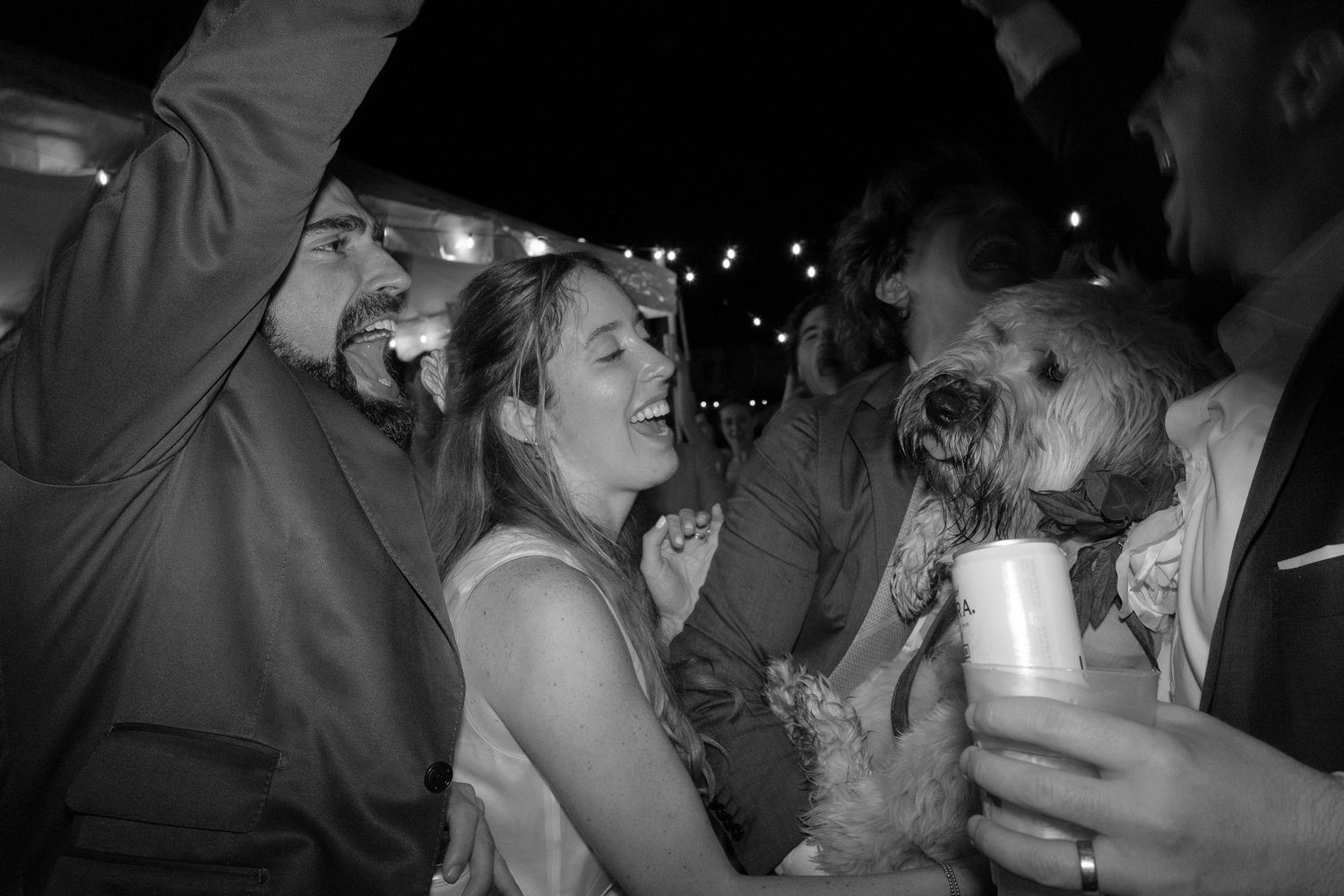 Black and white photo of laughing people and a fluffy dog at a festive outdoor party with string lights.