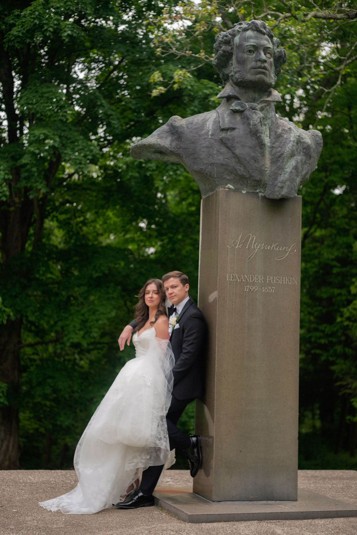 Bride and groom posing beside Alexander Pushkin bust monument in a lush green park on their wedding day.
