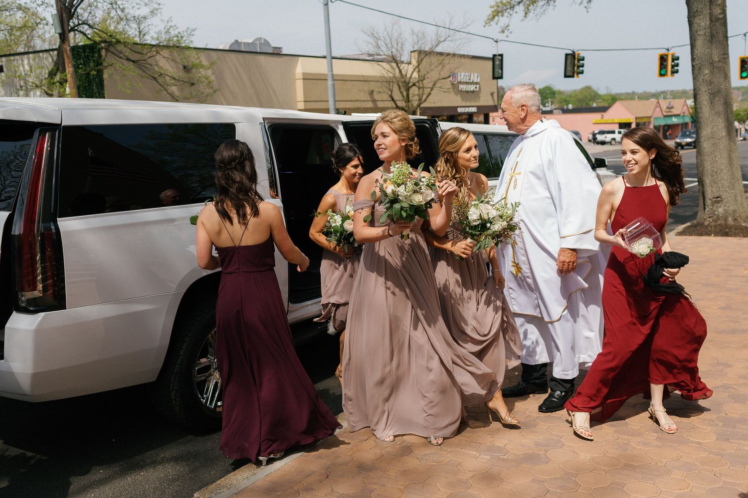 Bridesmaids in burgundy and taupe gowns holding floral bouquets walk toward a white limo with a priest outside a church.