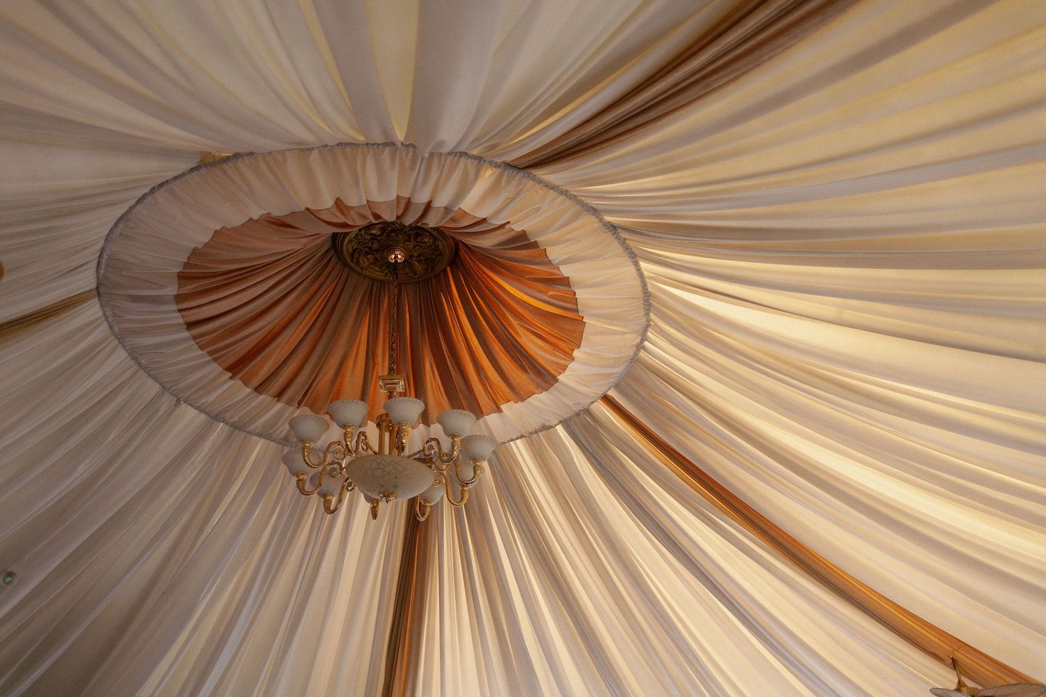 Interior view of an elegant marquee tent ceiling with draped white and gold fabric and a decorative chandelier centerpiece.