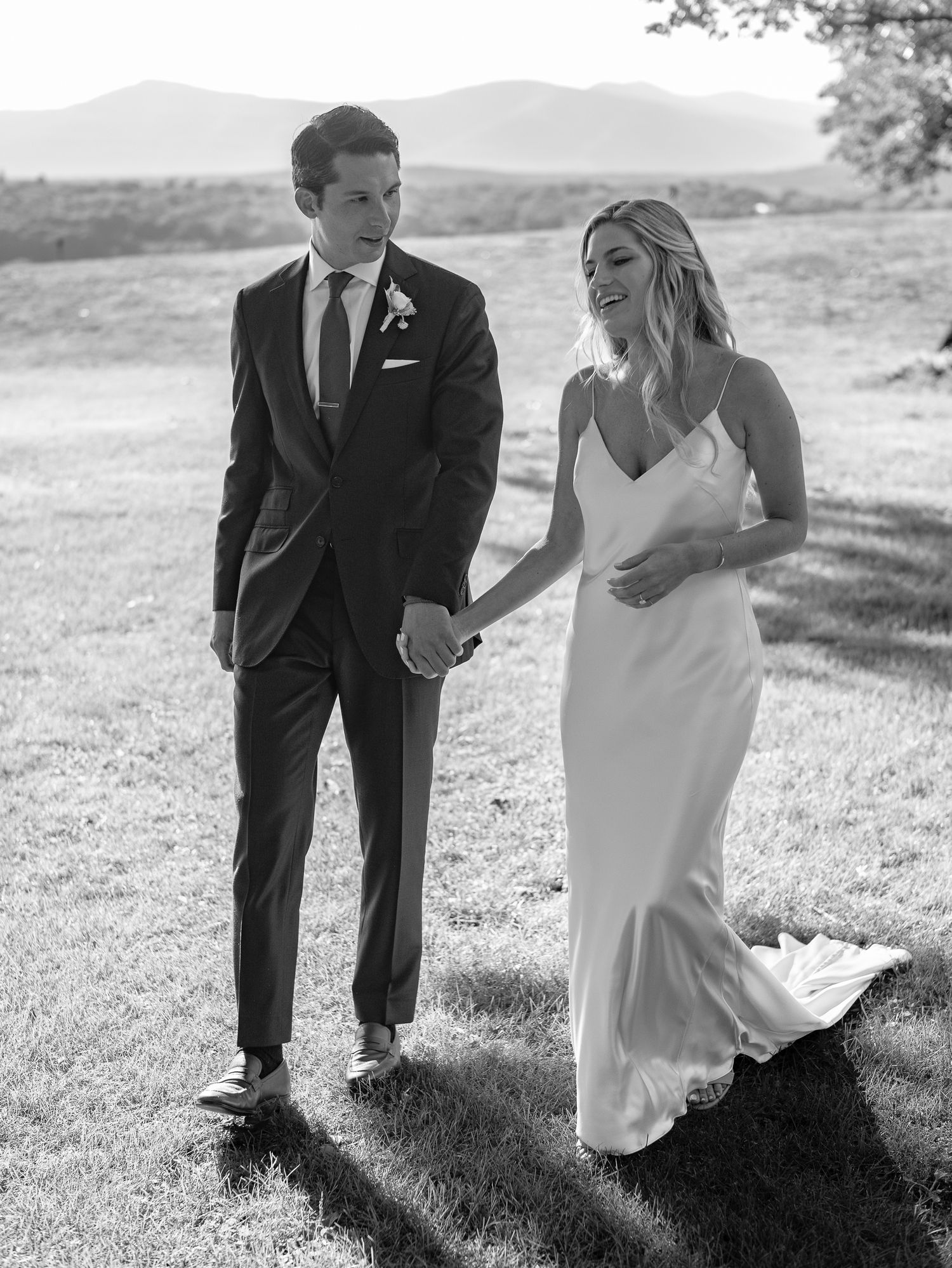 Black and white photo of a bride and groom holding hands walking outdoors with mountains in the background.