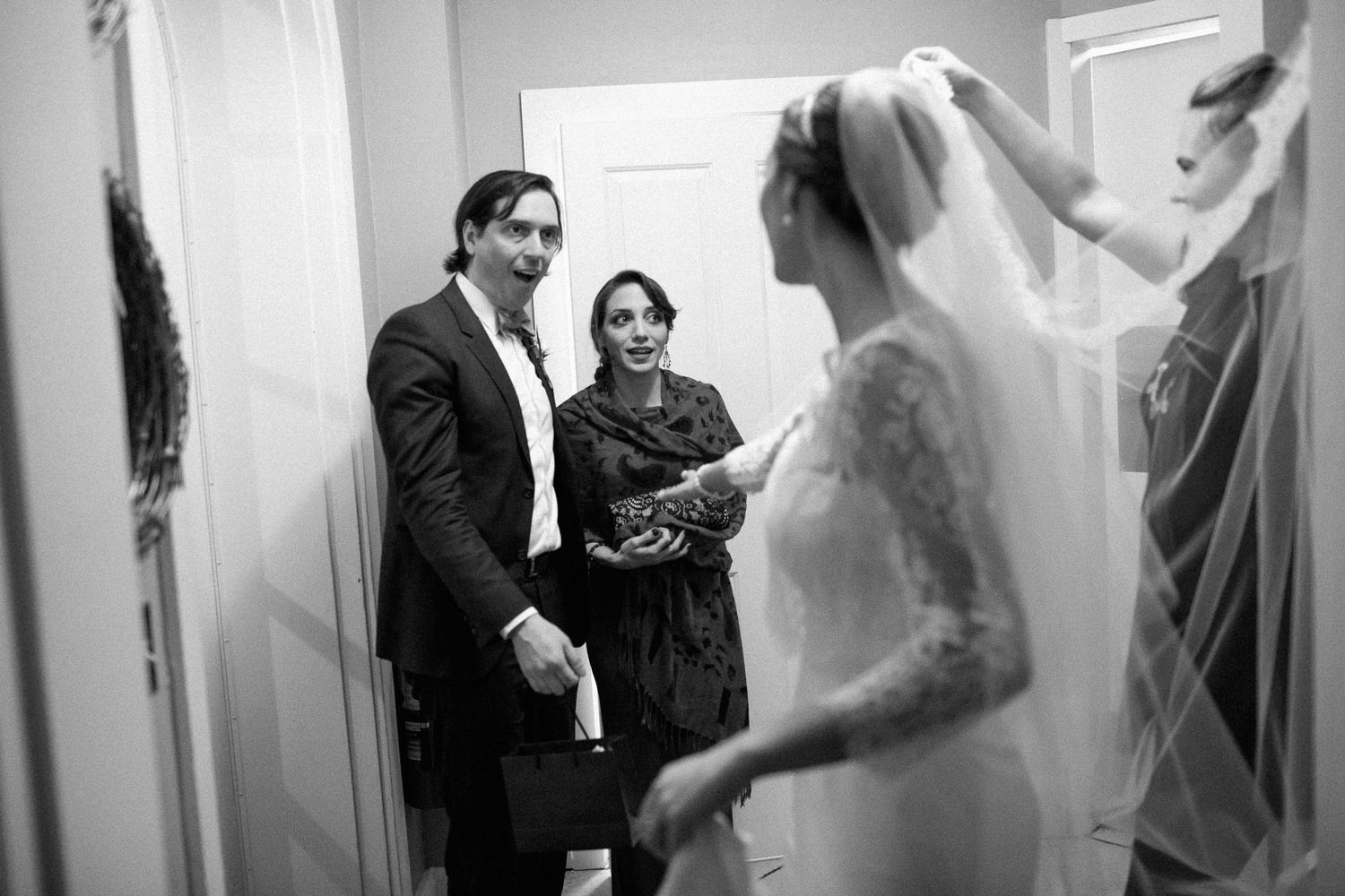 Black and white photo of a bride in a lace gown and veil being admired by guests in a hallway.