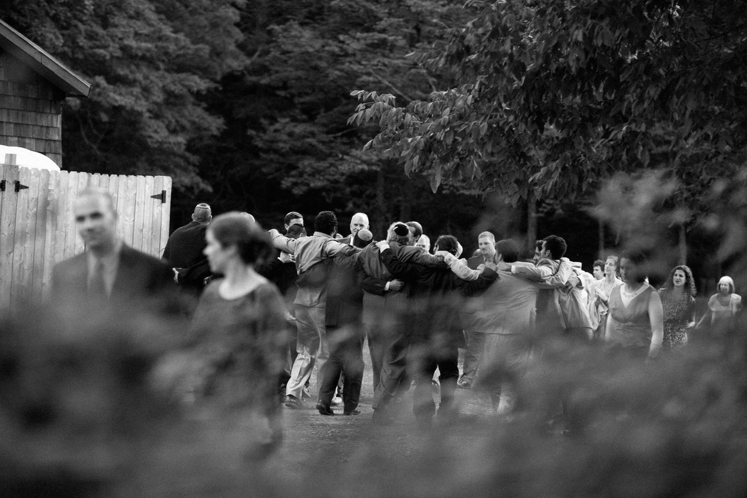 Black and white photo of a large group of people walking outdoors near trees and a wooden fence.
