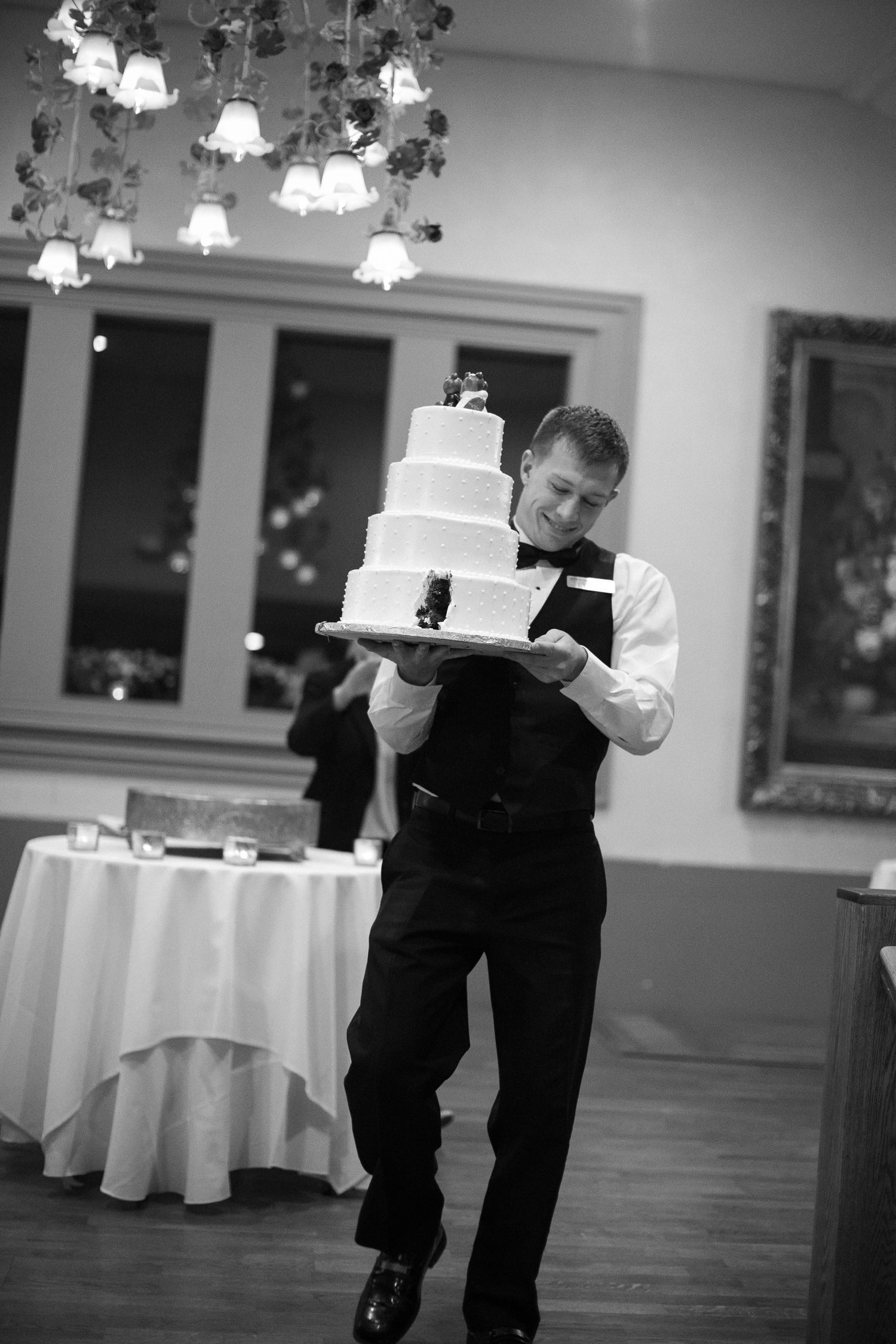 Waiter in bow tie carefully carries tiered white wedding cake across elegant reception hall in black and white photo.