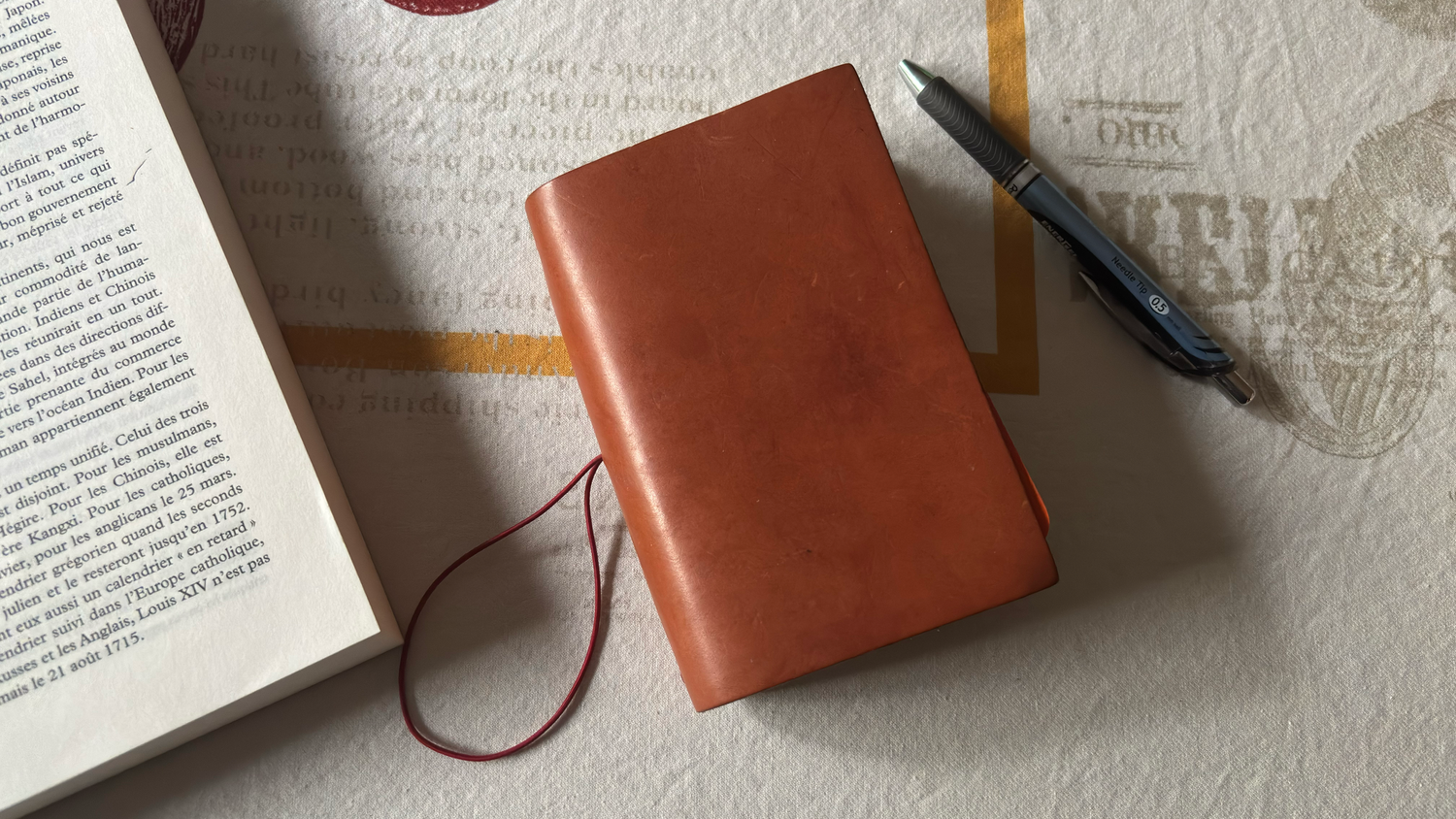 Tan leather journal cover with red cord bookmark beside an open book and blue pen on a white surface.