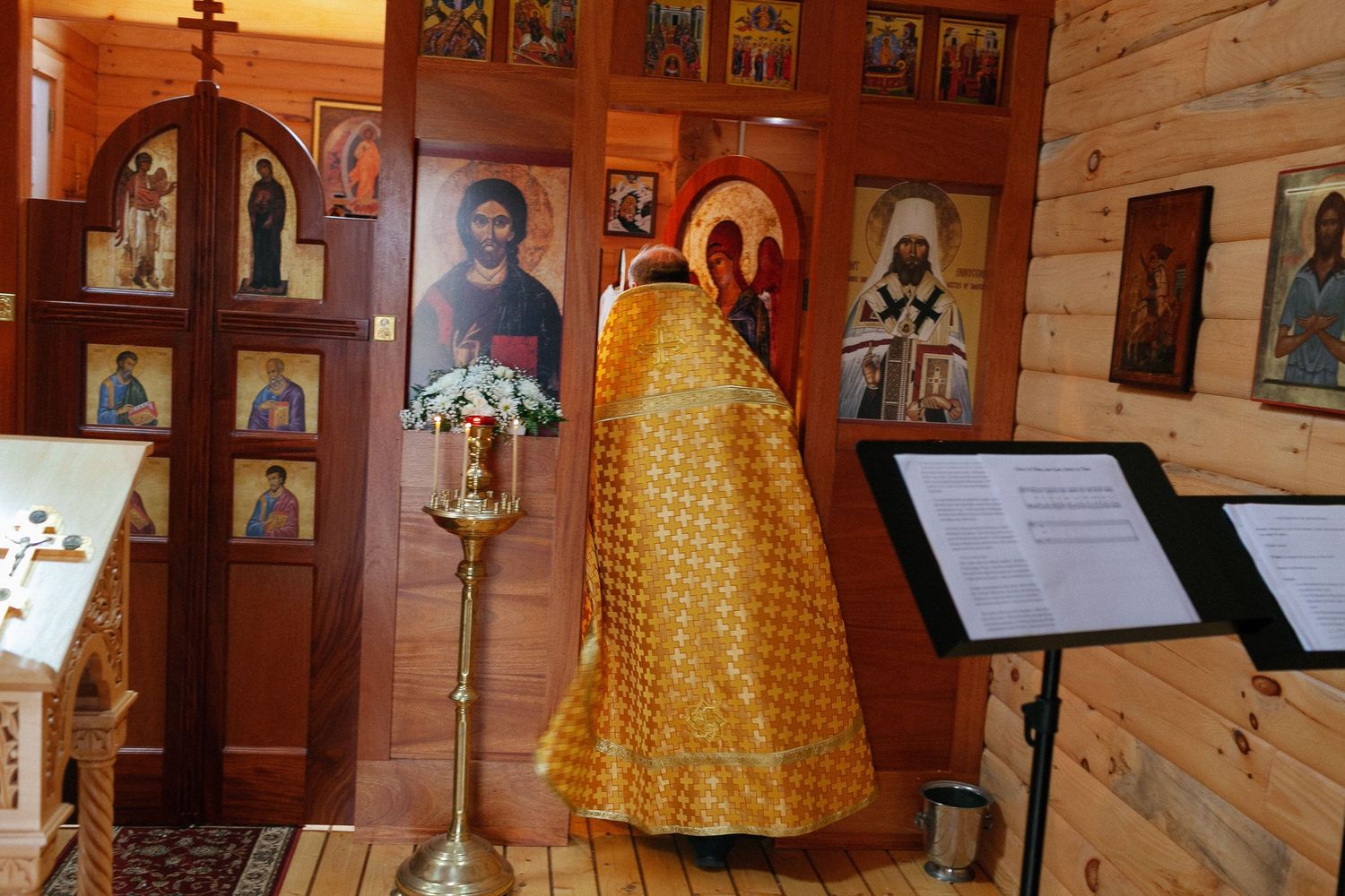 Orthodox priest in golden vestments stands before iconostasis in a wooden Russian Orthodox chapel with religious icons.