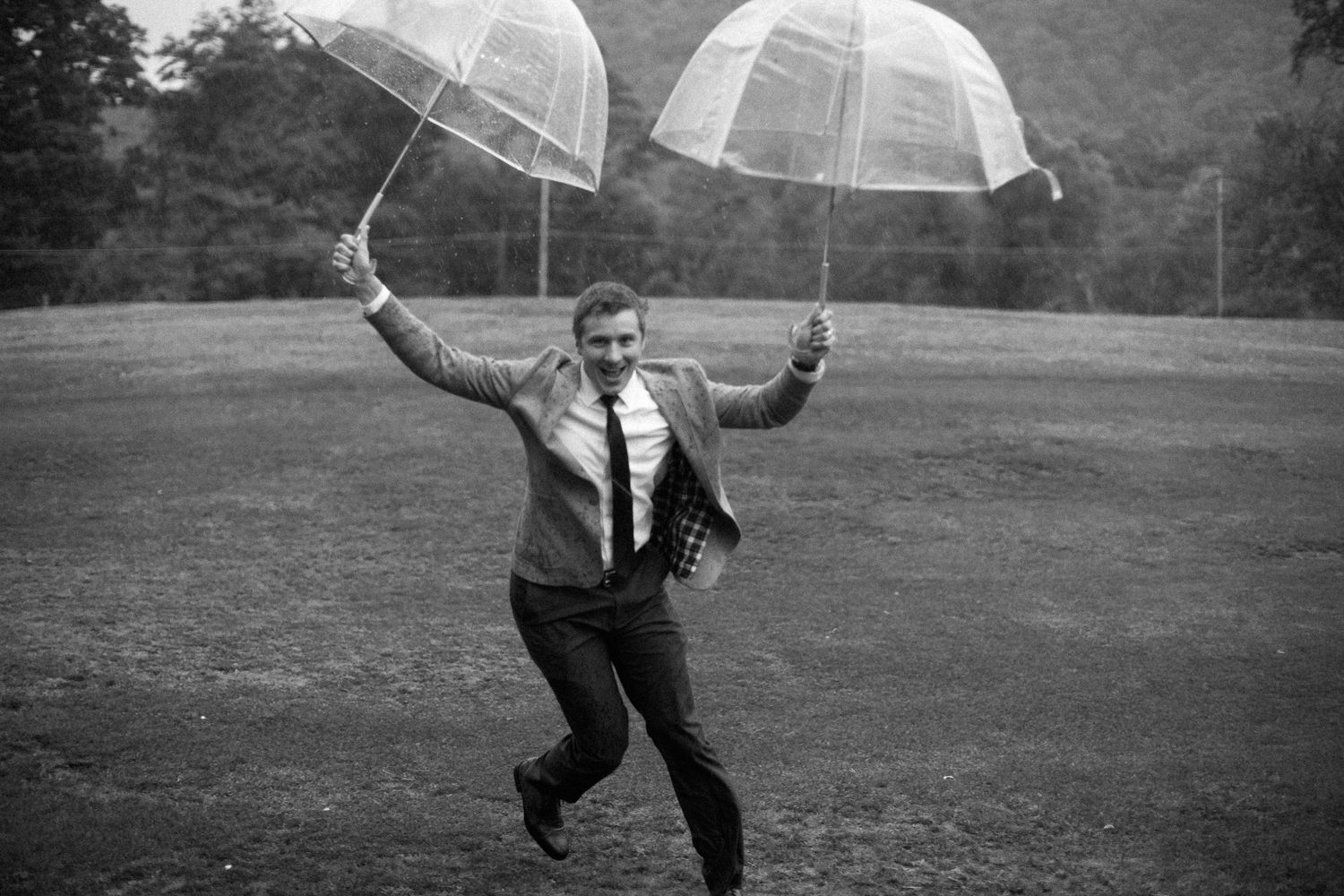Man in suit joyfully jumping in field holding two clear umbrellas in black and white photo.