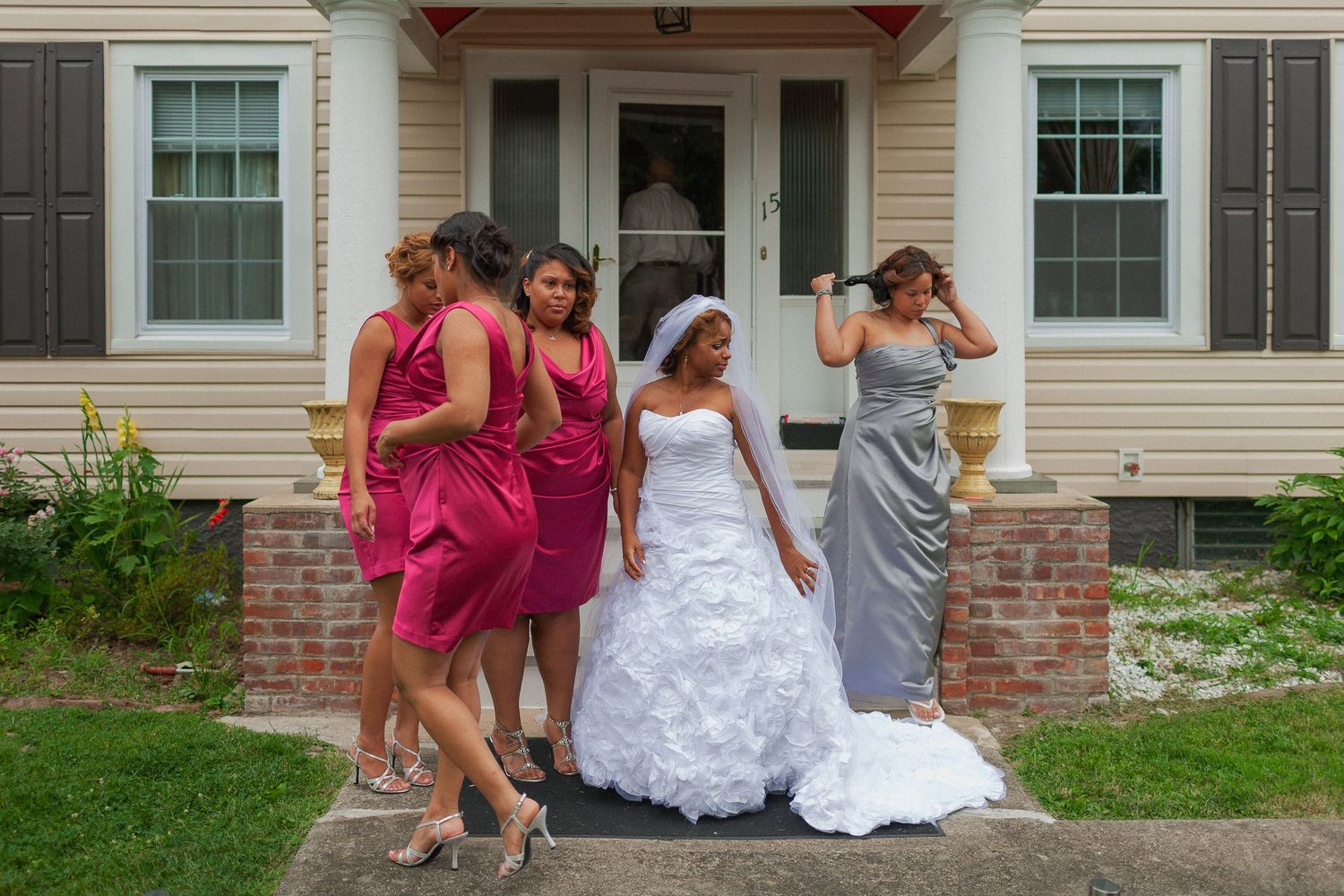 Bride in white gown poses with bridesmaids in pink and silver dresses outside a house on wedding day.