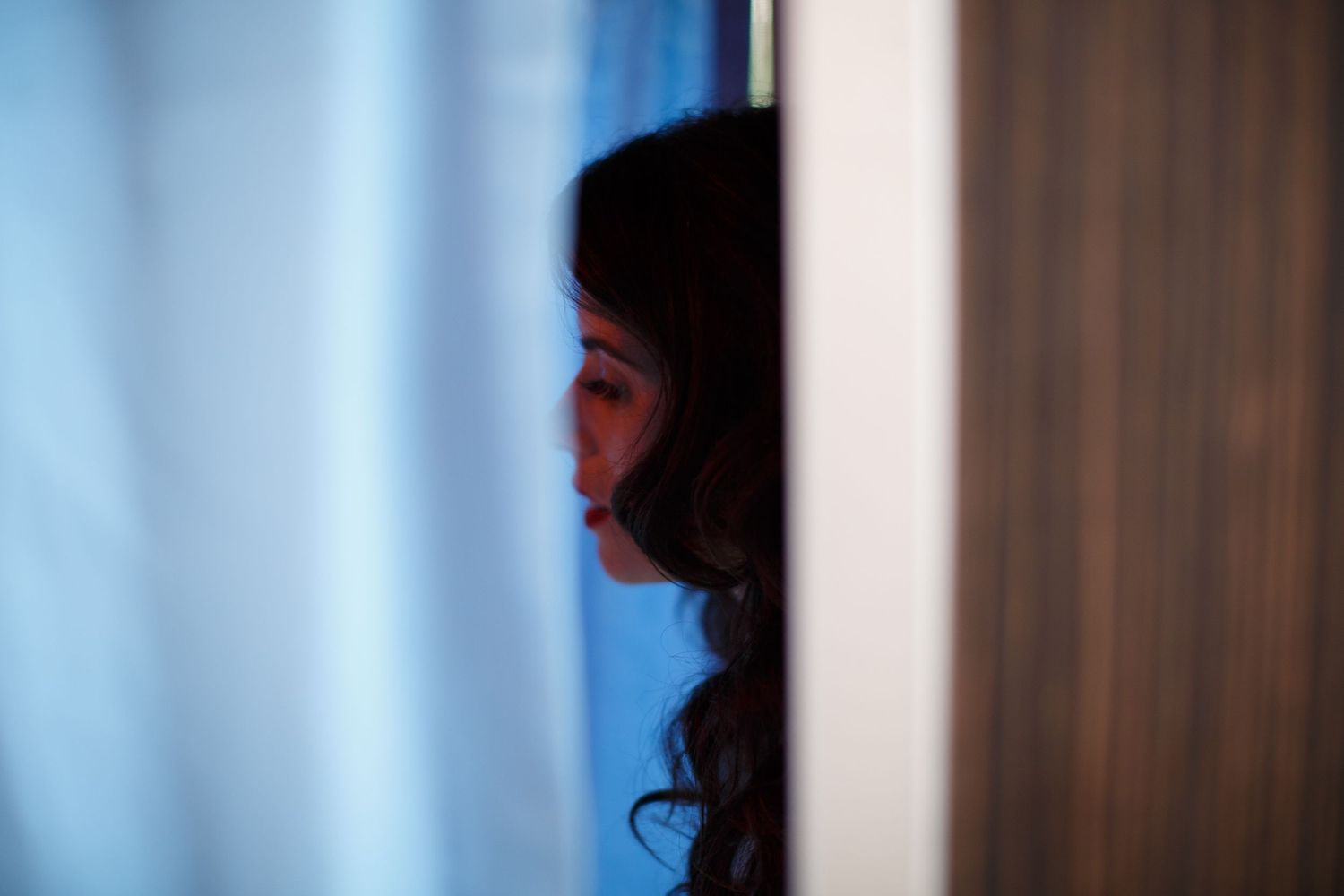 A woman with dark curly hair peers through a partially open door with blue-tinted curtains in the background.