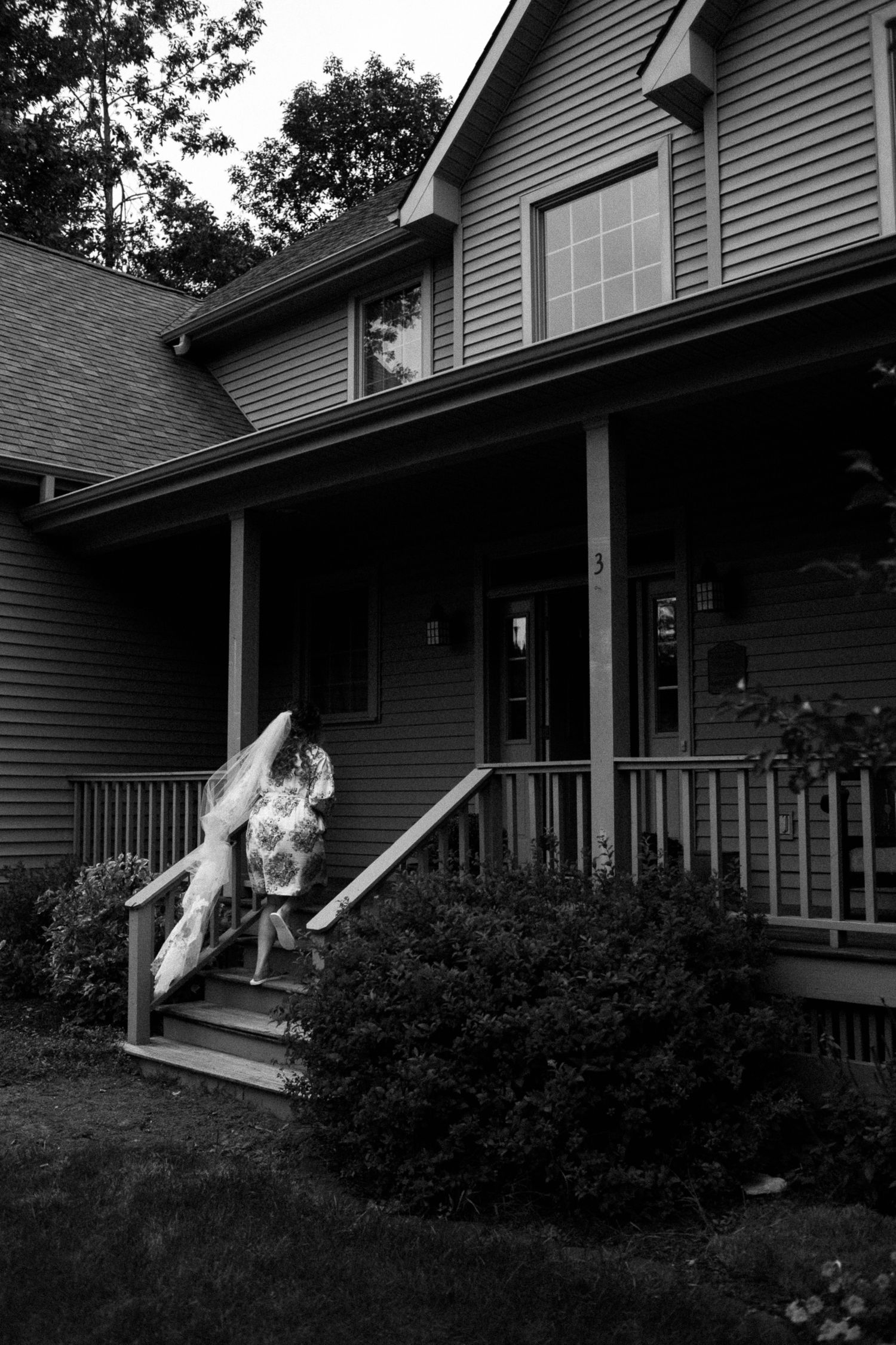 Black and white photo of a bride in a wedding dress carrying her gown up porch steps of a suburban two-story home.