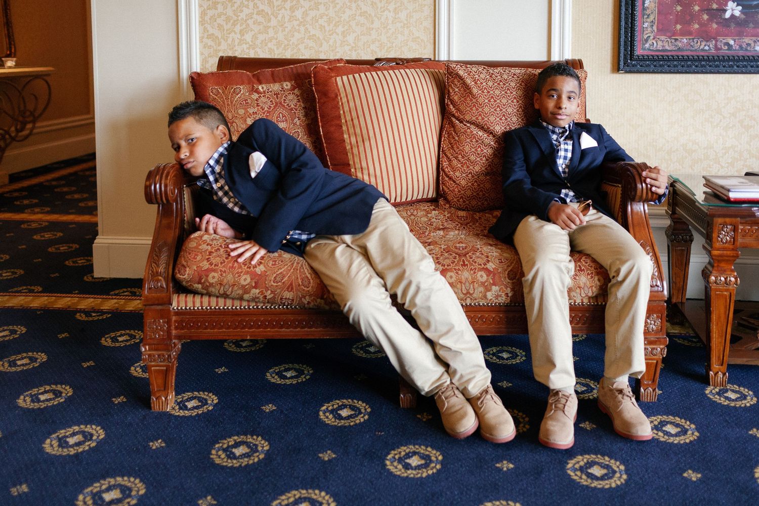 Two young boys in navy blazers and khaki pants sit on an ornate wooden sofa in an elegant room.