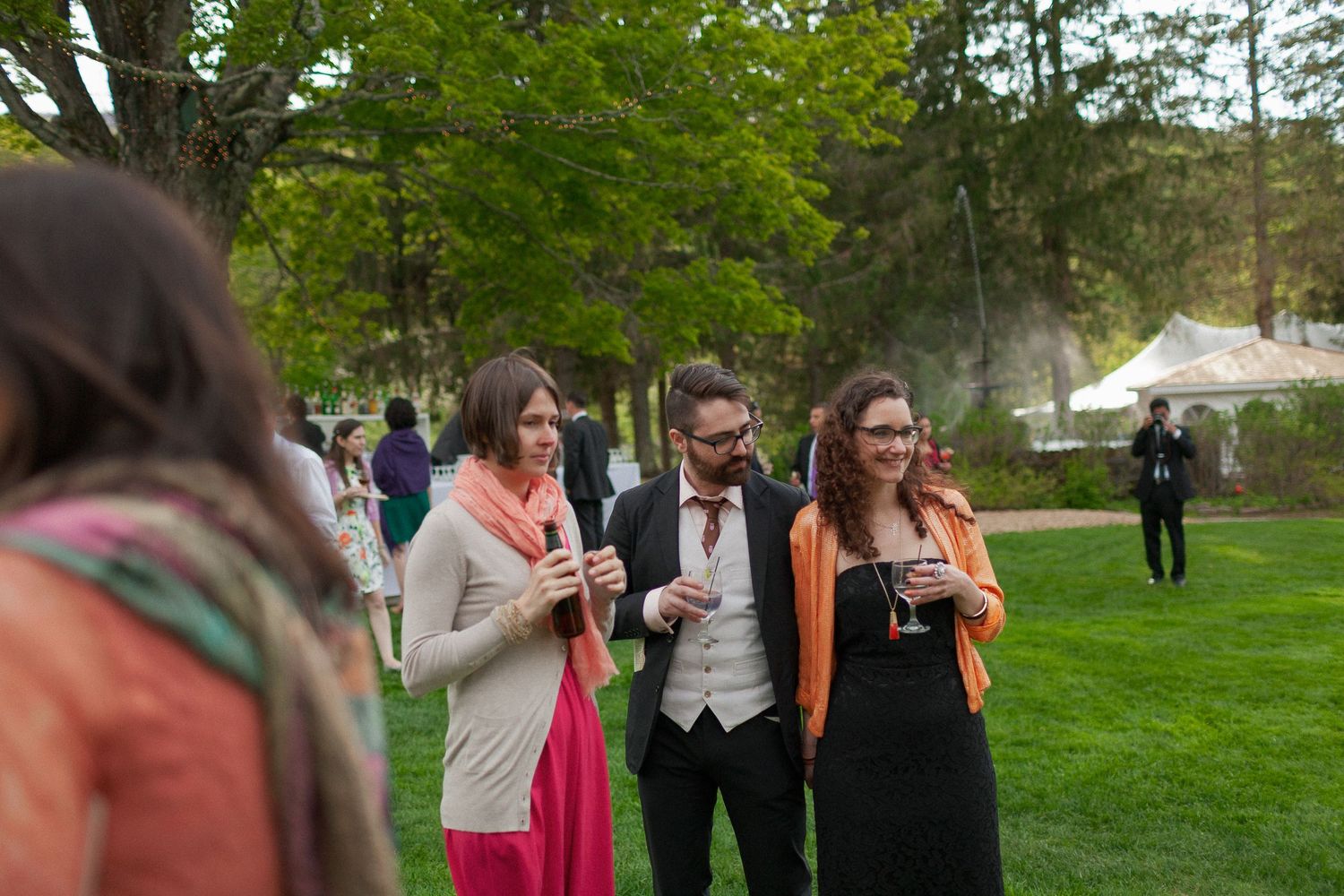 Guests socializing outdoors at an elegant garden party, holding drinks on a lush green lawn with trees in background.