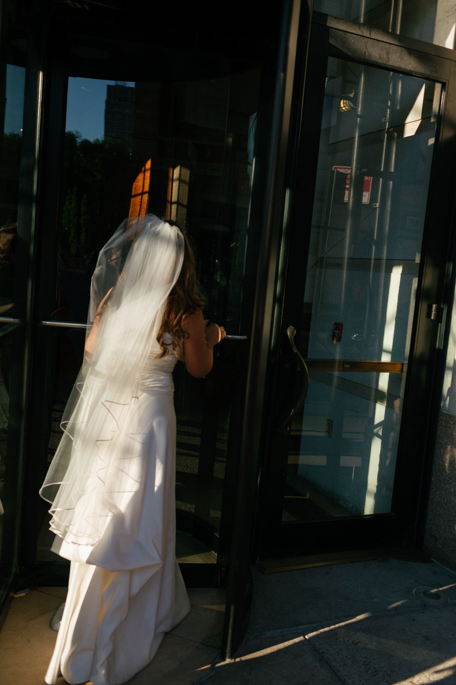Bride in white gown and veil opening glass door at wedding venue entrance.