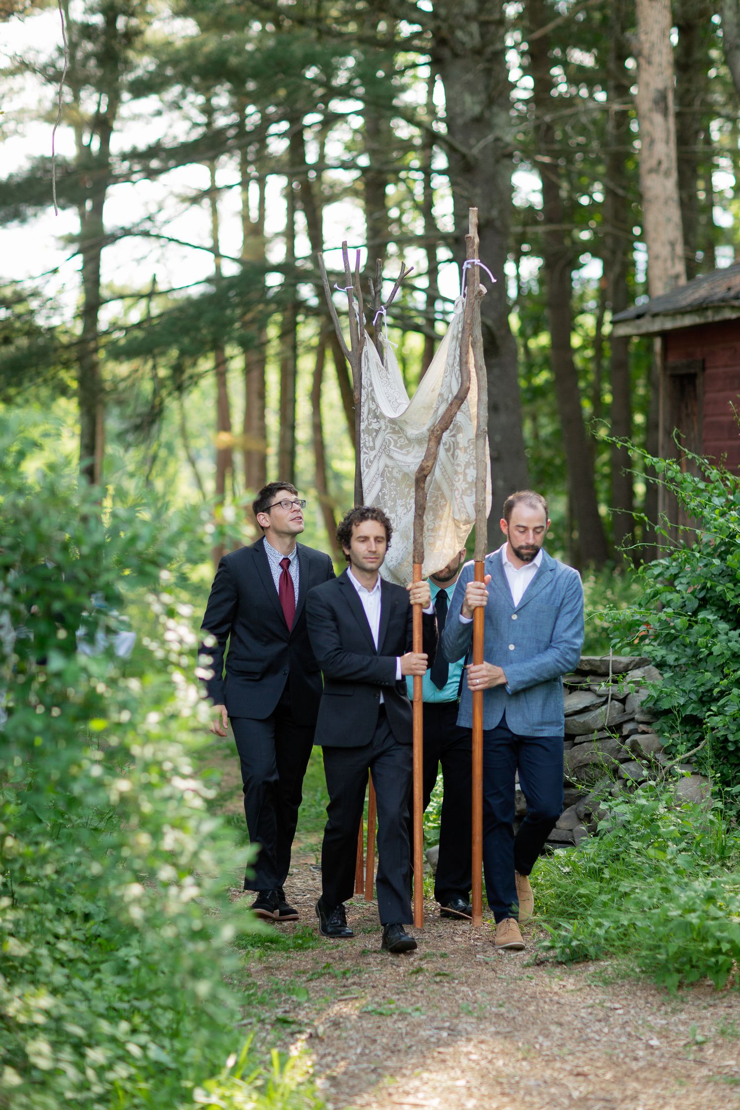 Three men in suits carry a decorative net installation along a wooded forest path near a rustic shed.
