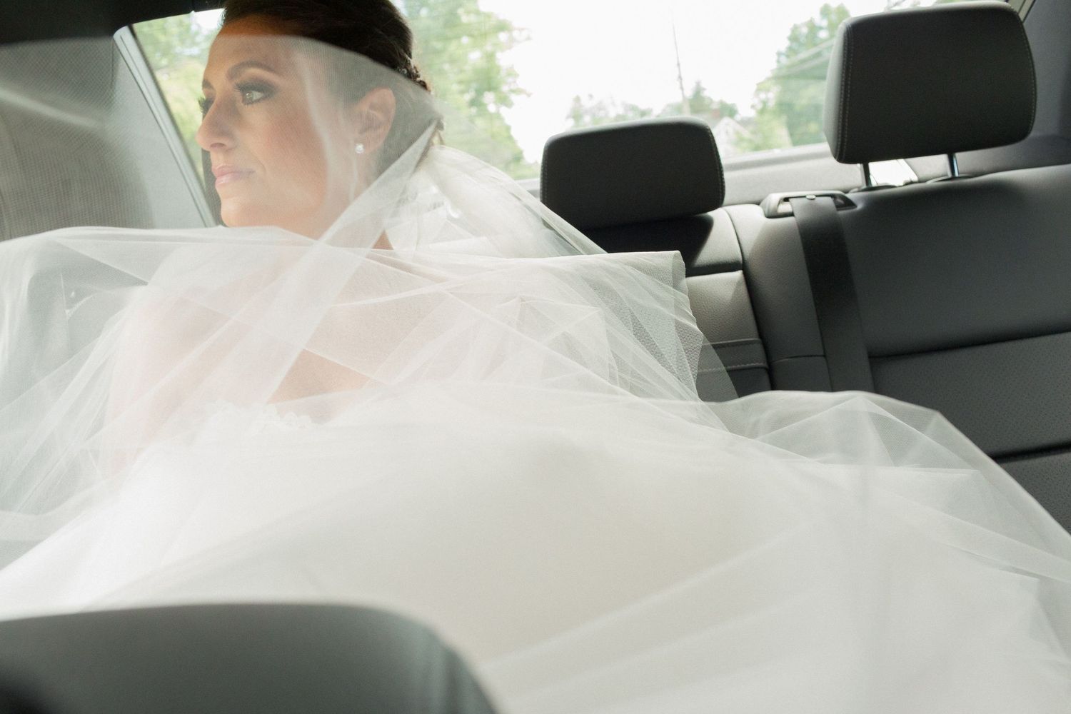 Bride in white wedding dress and veil sitting in back seat of car on wedding day.