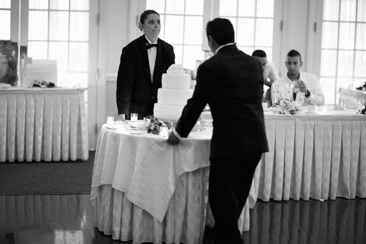 Two men in tuxedos stand near a white wedding cake on a draped table at an elegant reception venue.