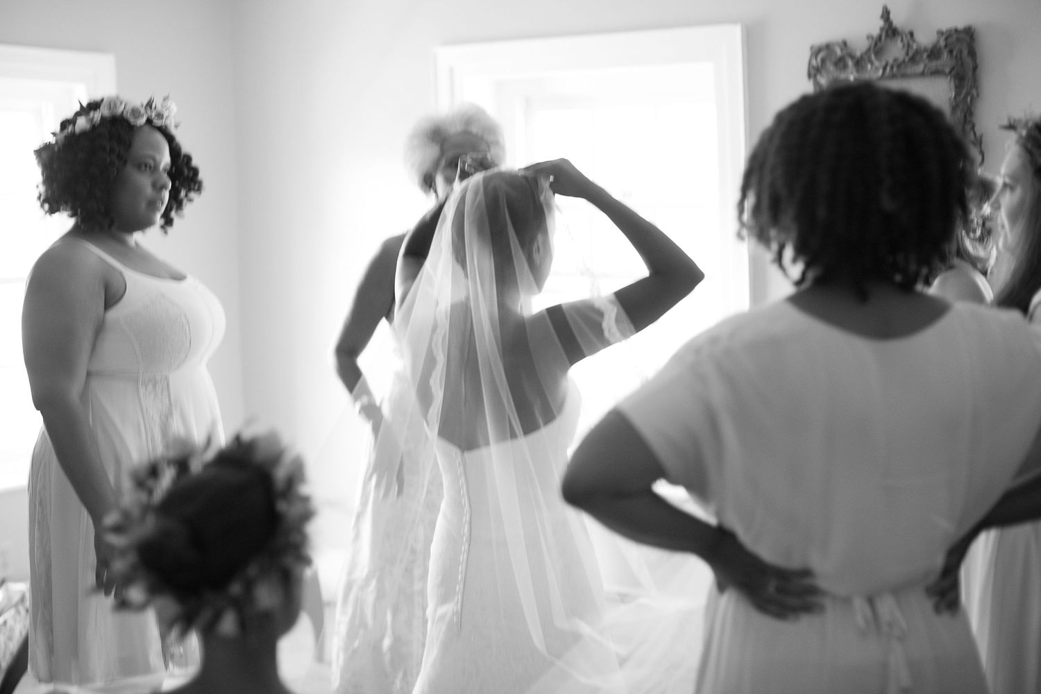 Black and white photo of a bride adjusting her veil near a window, surrounded by bridesmaids in white dresses.
