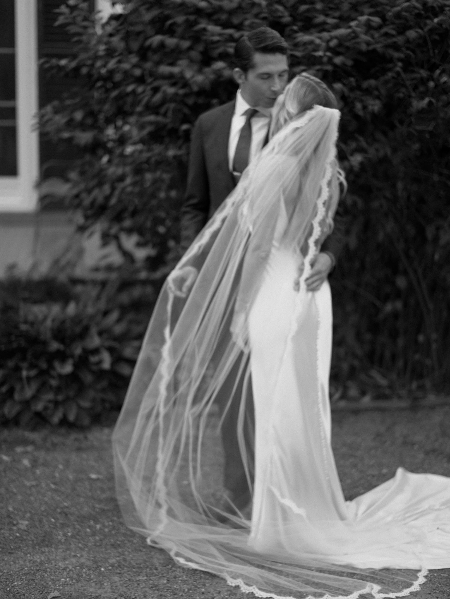 A black and white wedding photograph showing a flowing veil caught in the breeze during an outdoor portrait session.