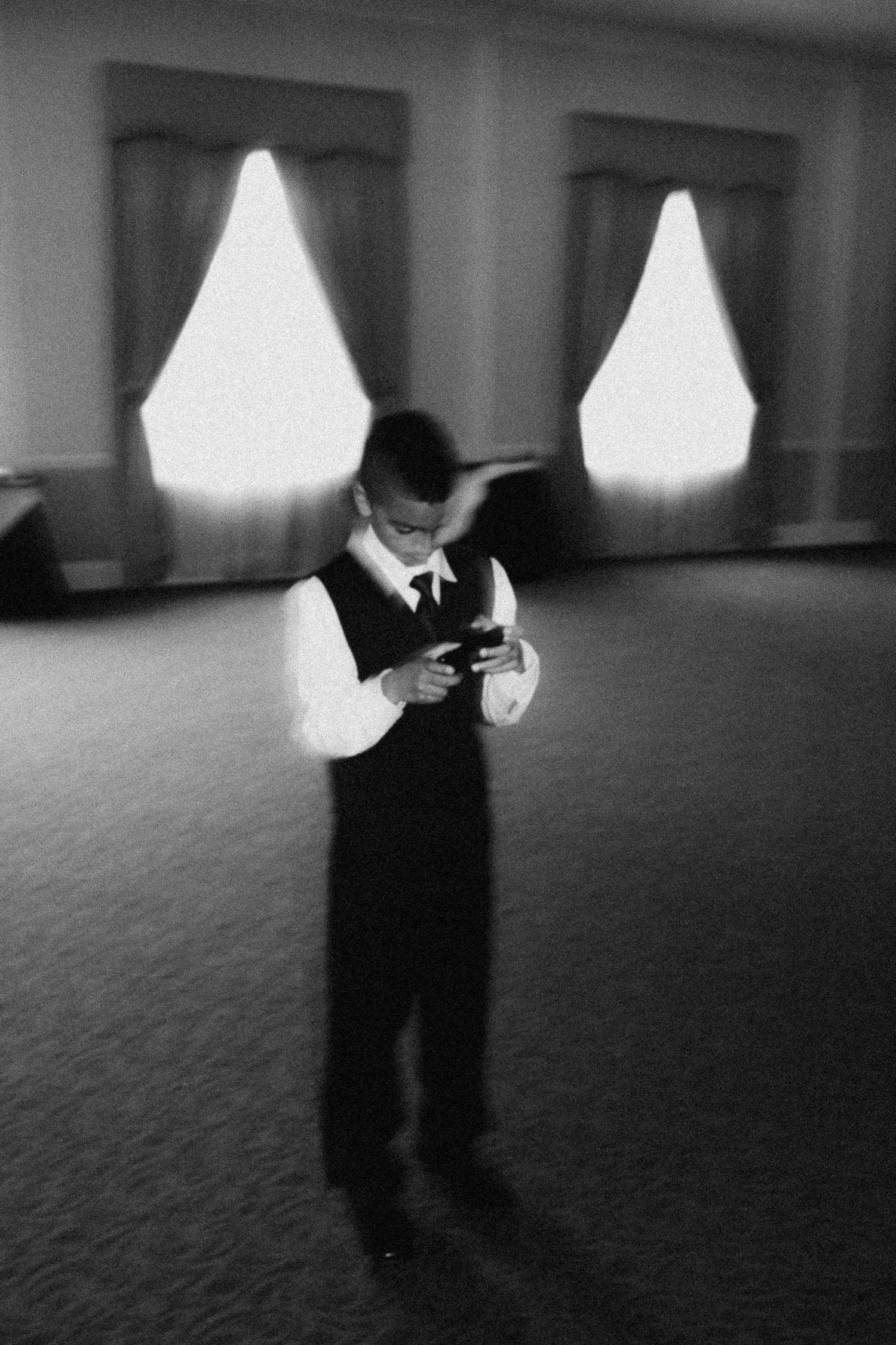 Black and white photo of a boy in formal vest checking his phone in an elegant ballroom with draped windows.