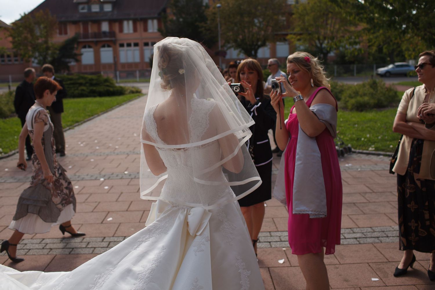 Bride in white wedding gown and veil surrounded by guests outdoors at a sunny wedding ceremony.