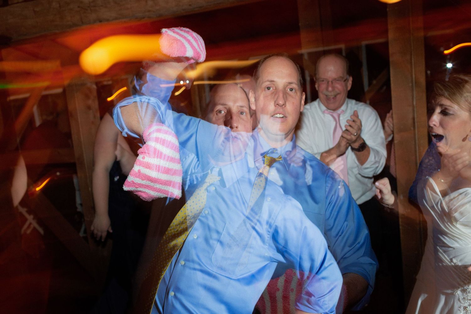 Guests dancing energetically at a wedding reception, with a man in a blue shirt being playfully spun on the dance floor.