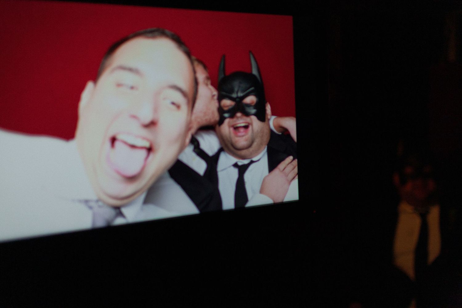 Two men in suits pose on screen, one wearing a Batman mask, making funny faces against a red background.