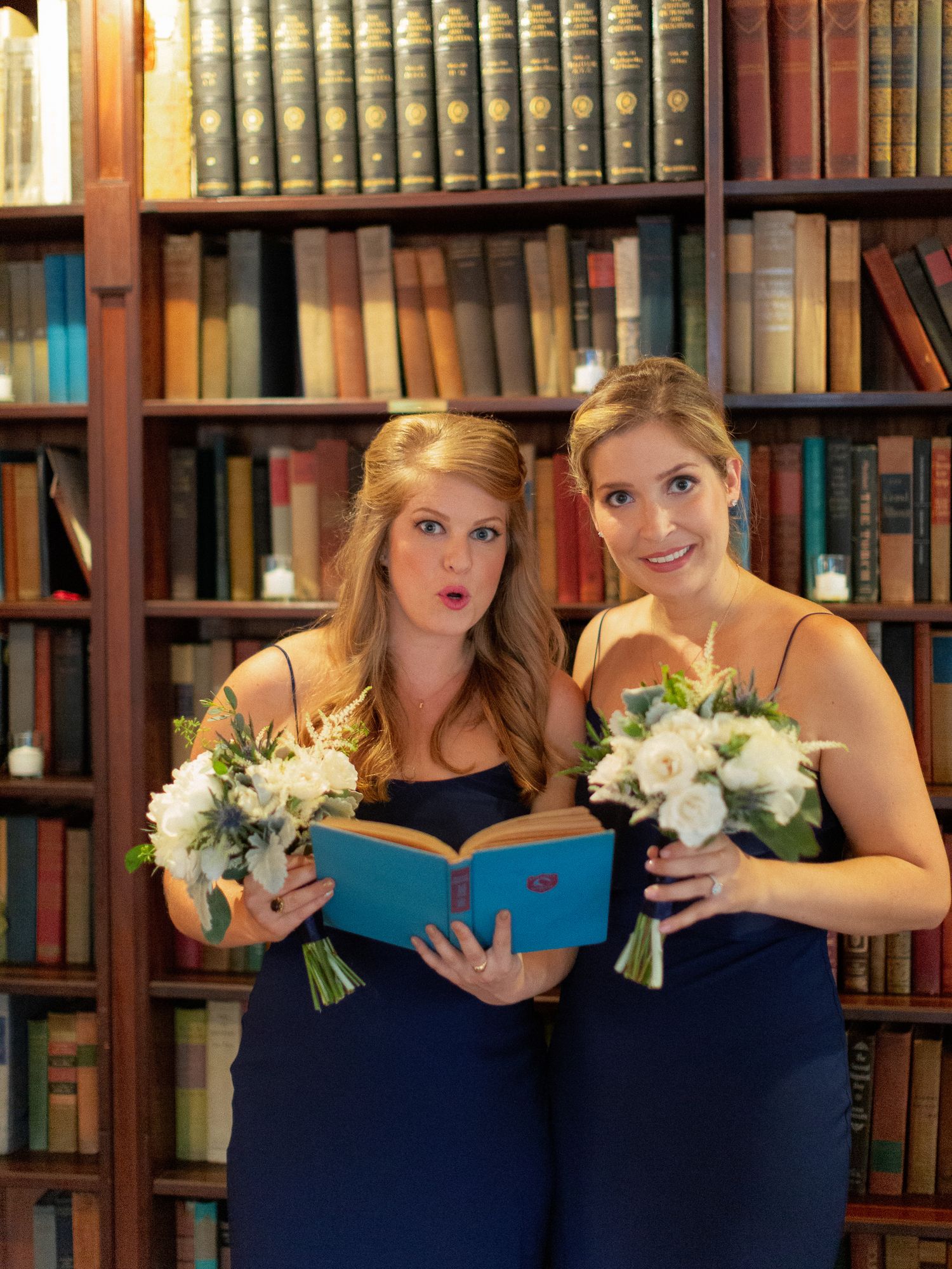 Two bridesmaids in navy dresses holding white bouquets pose in front of a vintage library bookshelf.