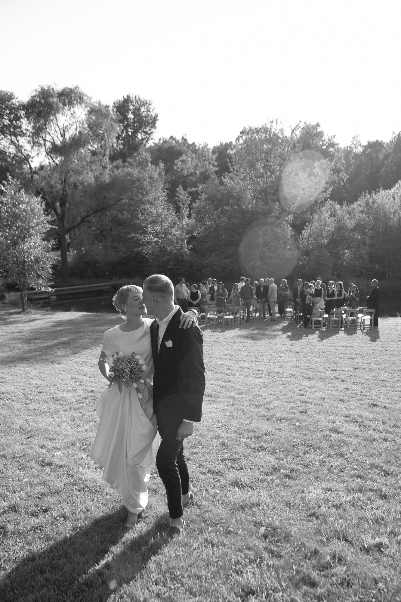 A couple walks together on a grassy hillside during an outdoor wedding ceremony with guests in the background.