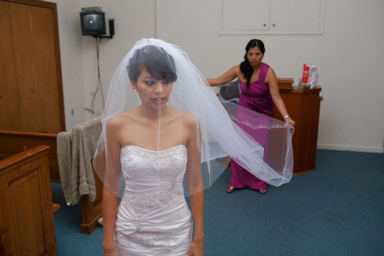Bride in white strapless gown and veil with bridesmaid in purple dress adjusting veil before wedding ceremony.