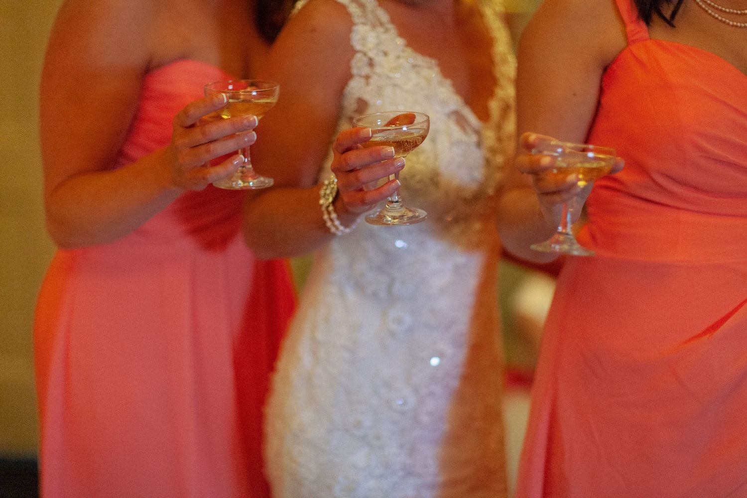 Bride in white lace dress and bridesmaids in coral gowns holding martini glasses at wedding reception.