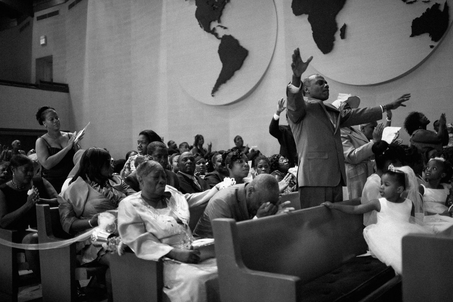 Black and white photo of congregation raising hands in worship inside a church with a world map mural.