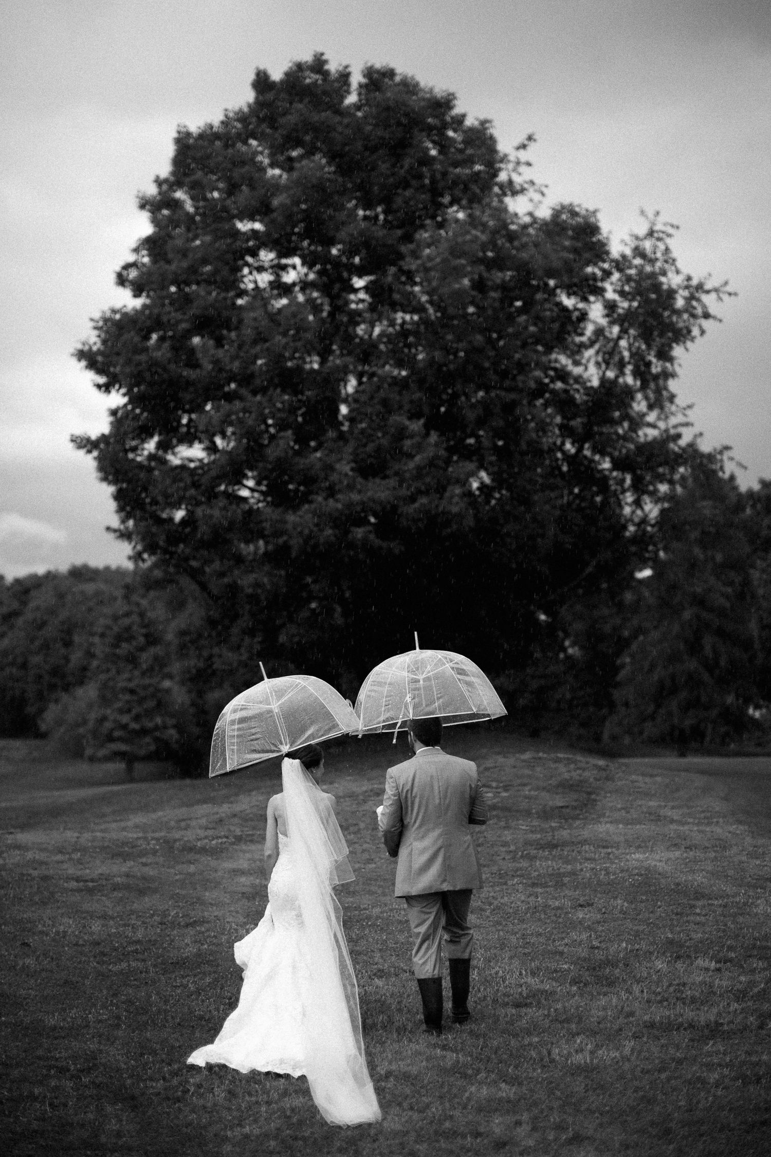 Black and white photo of bride and groom walking with clear dome umbrellas on a rainy wedding day.